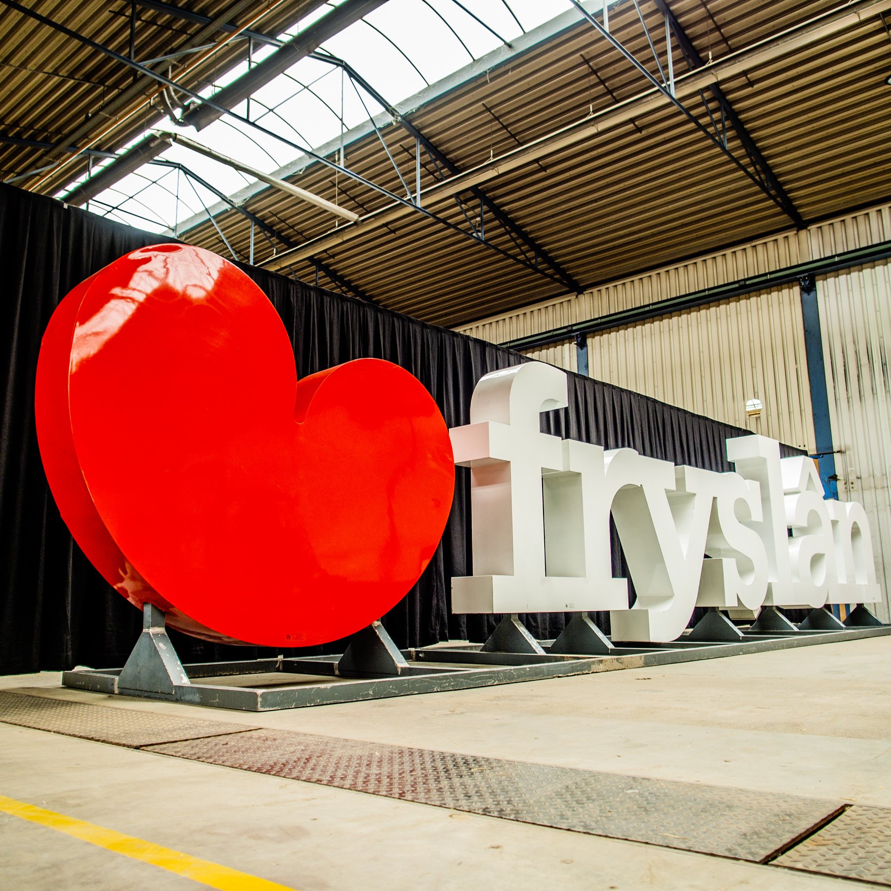 Een grote rode hart en witte letters die samen 'Liefde Lys' vormen, geplaatst binnen een industrie-achtige ruimte met een metalen plafond en zwarte zijwand.
