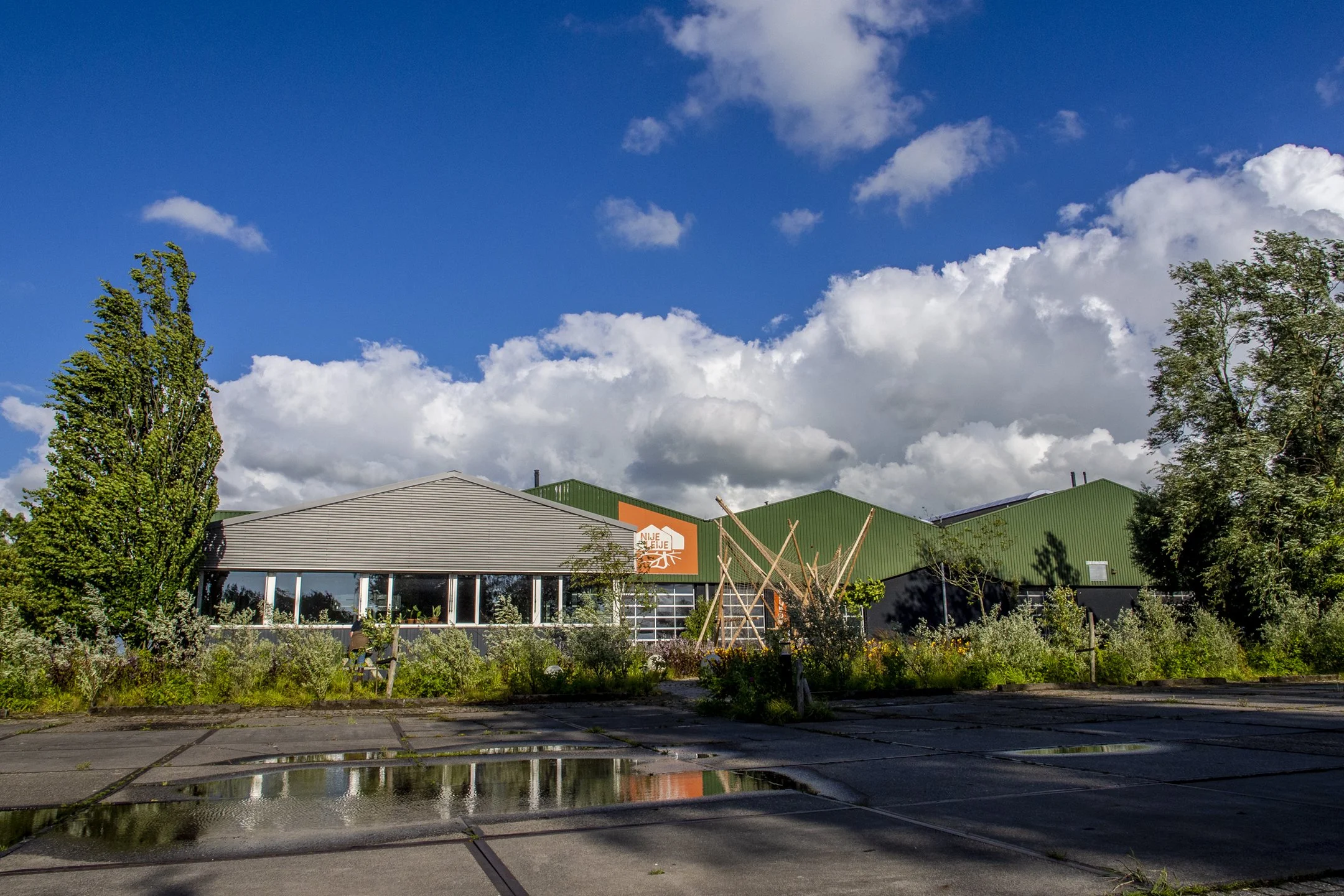 Een industrieel gebouw met een grijze en groene metalen dak, omgeven door bomen en planten, onder een bewolkte blauwe lucht. Nijeleije Friesland Eventlokatie