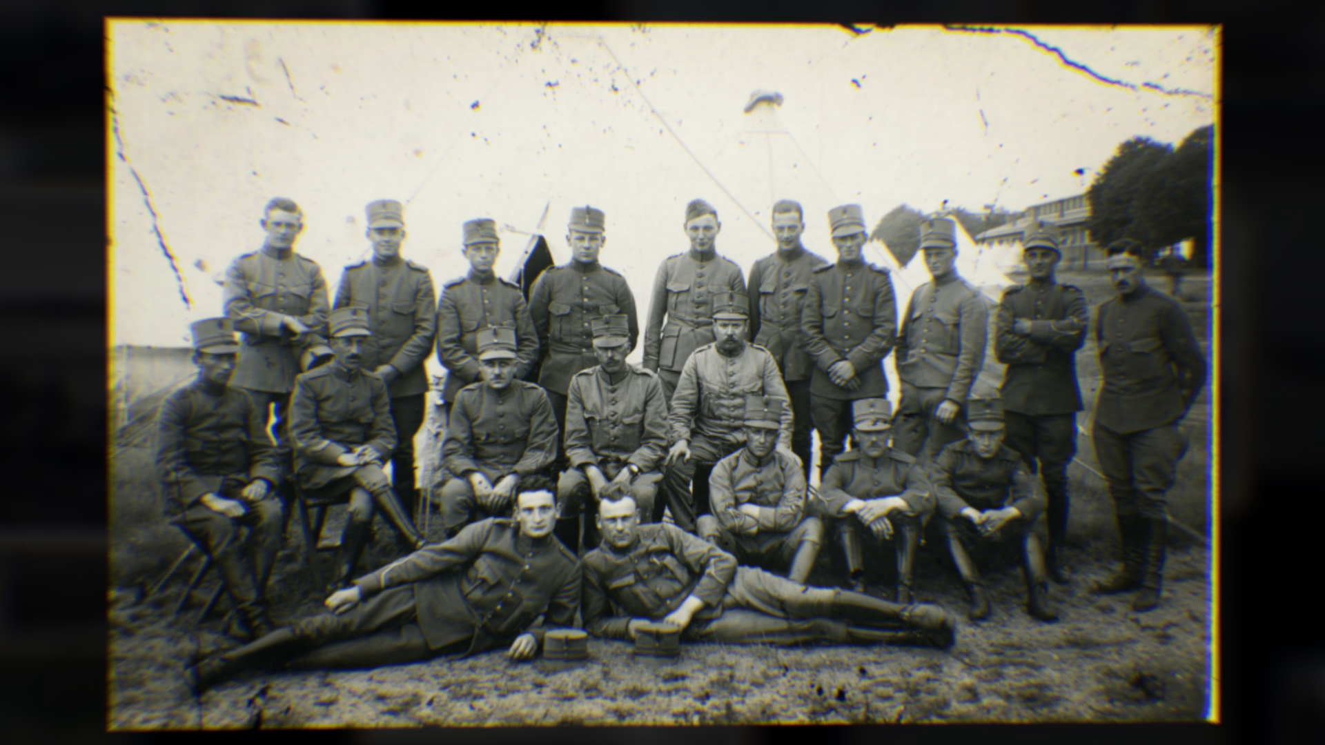 Zwart-wit foto van een groep mannen in militaire uniformen, zittend en staand in rij. Ze dragen epaulette en mutsen, met een open veld en enkele tenten op de achtergrond.