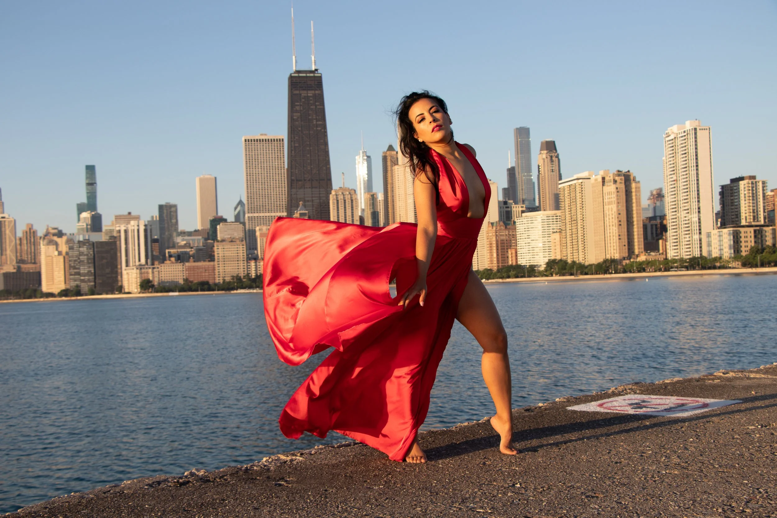 A woman in a red dress standing on a rocky shoreline near a body of water with a city skyline in the background, including tall skyscrapers.