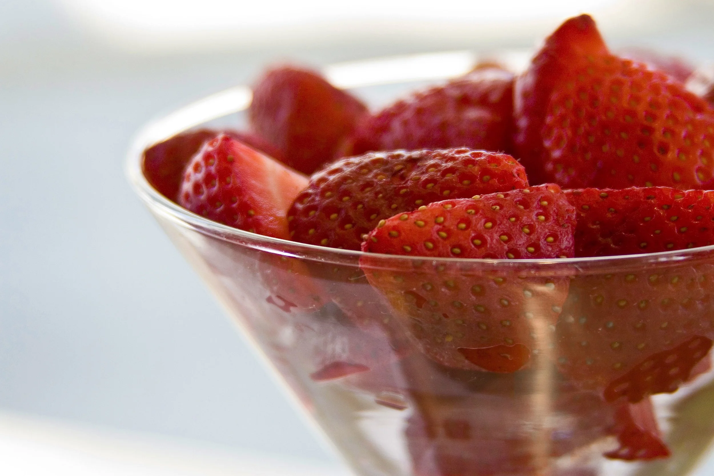 Close-up of a glass bowl filled with fresh strawberries, some sliced and some whole, on a white surface.