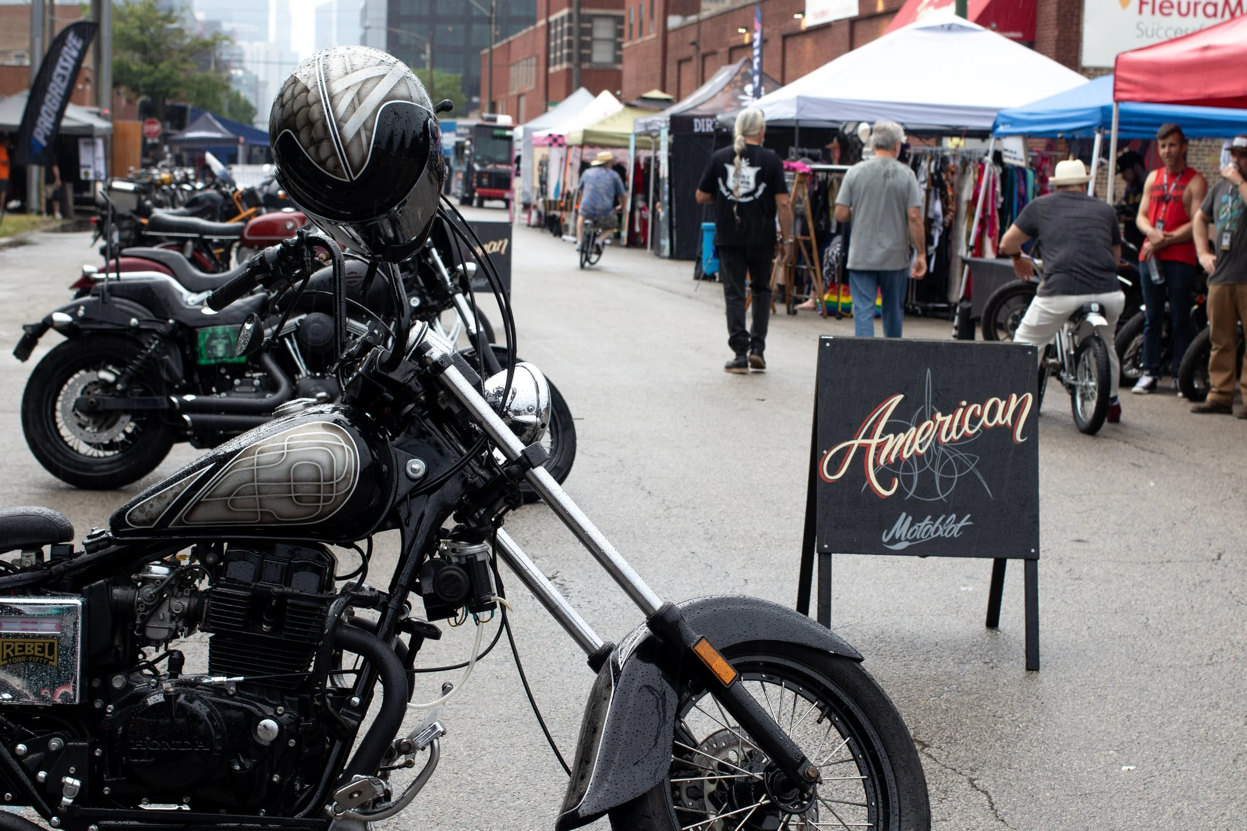 Close-up of a black motorcycle with a helmet hanging on the handlebars, parked on a city street during a street fair with tents, people walking, and vendor signs visible in the background.