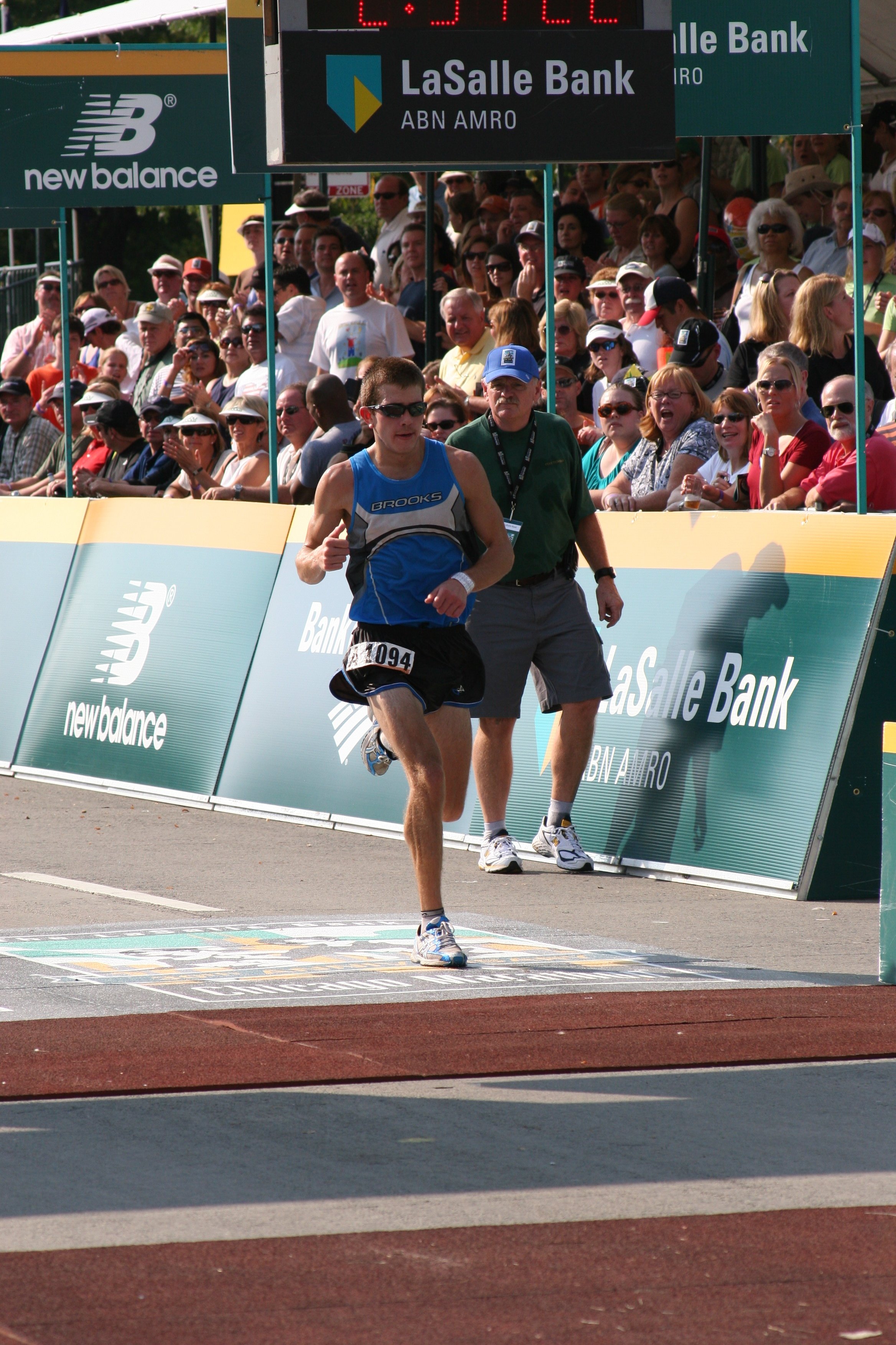 A male runner in a blue tank top and shorts crossing a finish line with a crowd of spectators cheering behind a barrier. The event takes place outdoors on a sunny day.