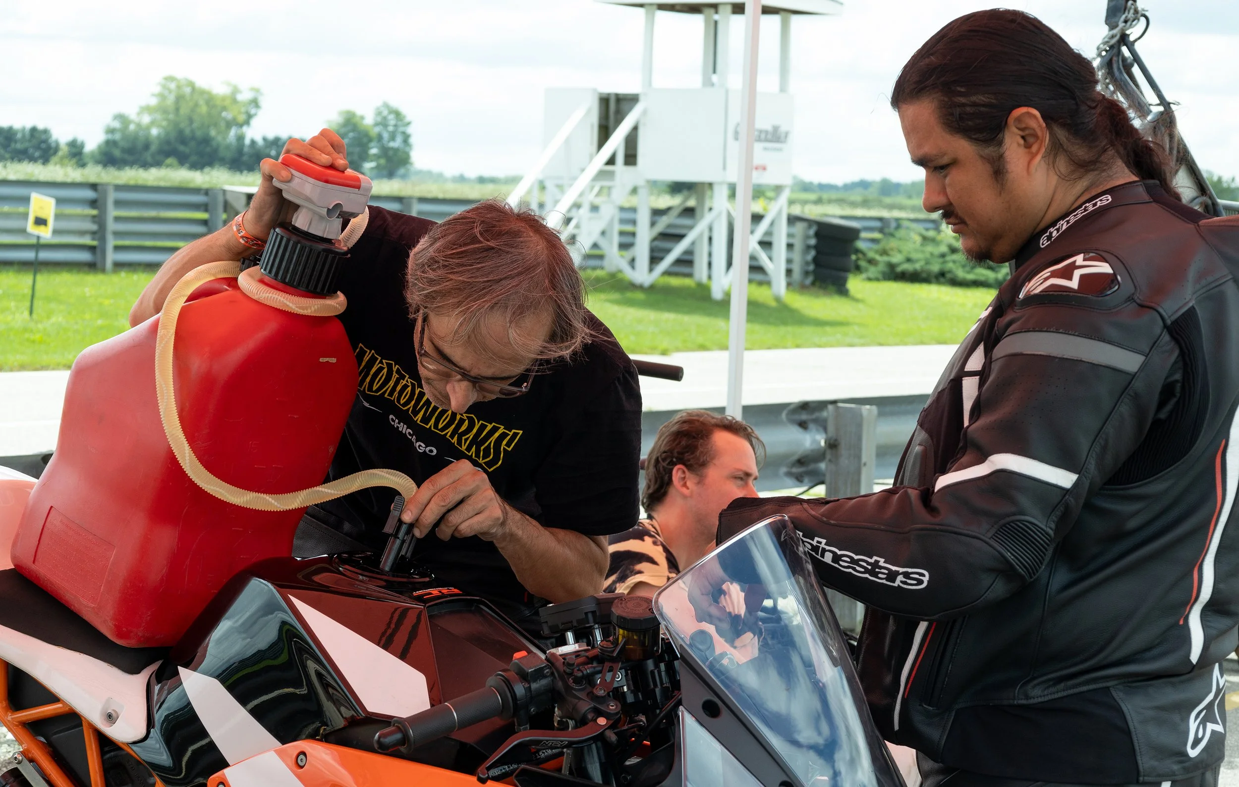 Two men, one elderly and one younger, working on a motorcycle at an outdoor event, with a person seated nearby and standing in front of a white structure and greenery in the background.