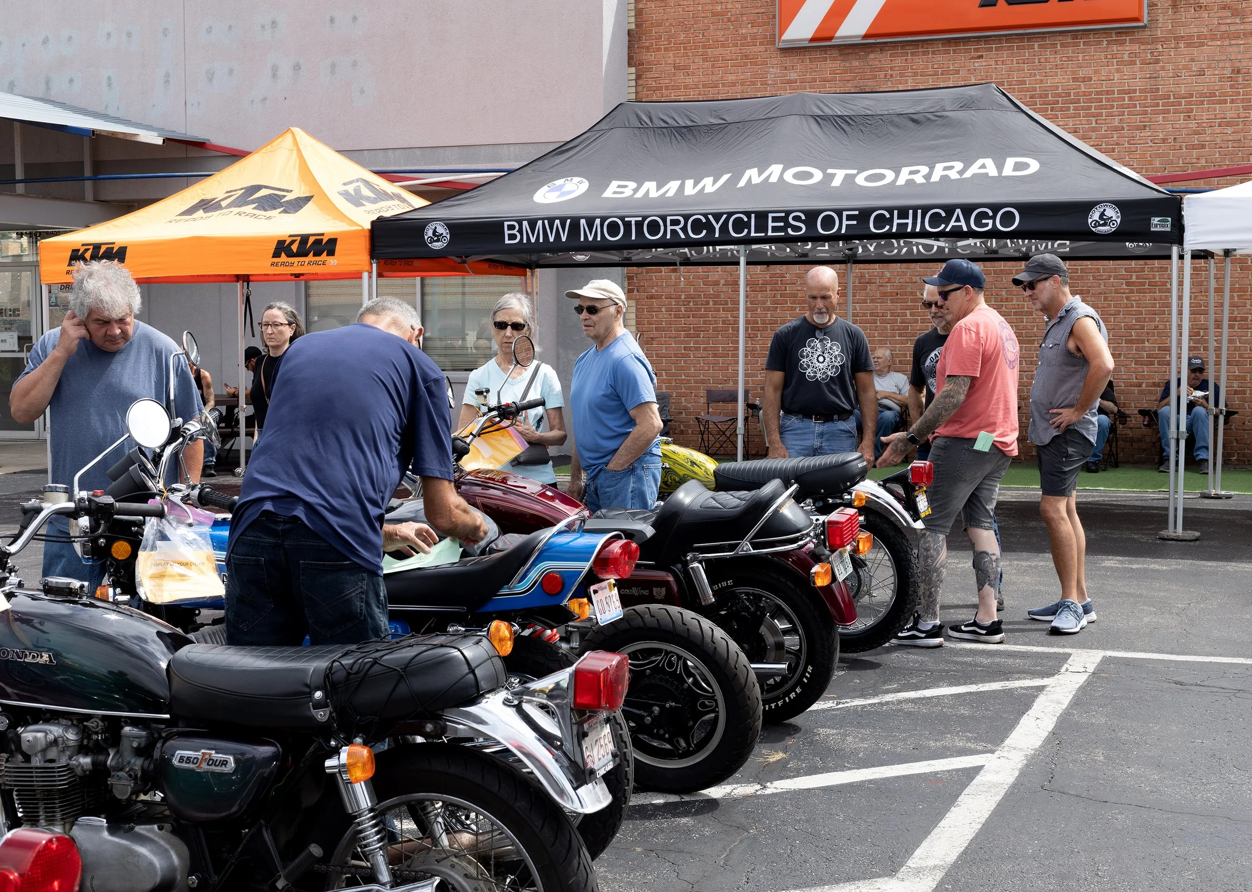 A group of people gathered around classic motorcycles under a BMW Motorrad tent at an outdoor event. There are multiple motorcycles parked, and some people are inspecting or talking about the bikes. The scene is set in a parking lot with a brick buil