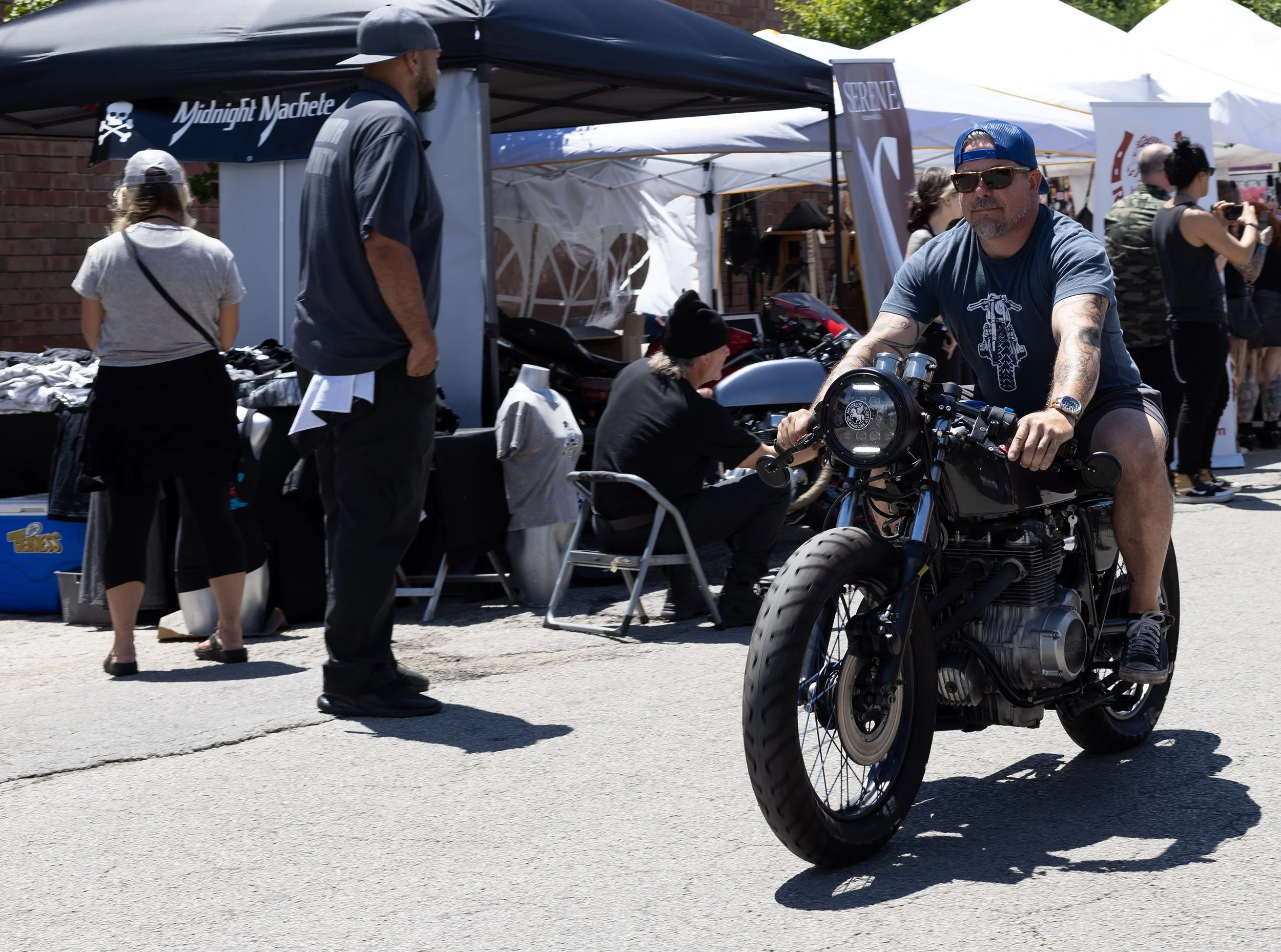 A man with tattoos and a blue cap riding a black motorcycle at an outdoor event, with tents and other people in the background.