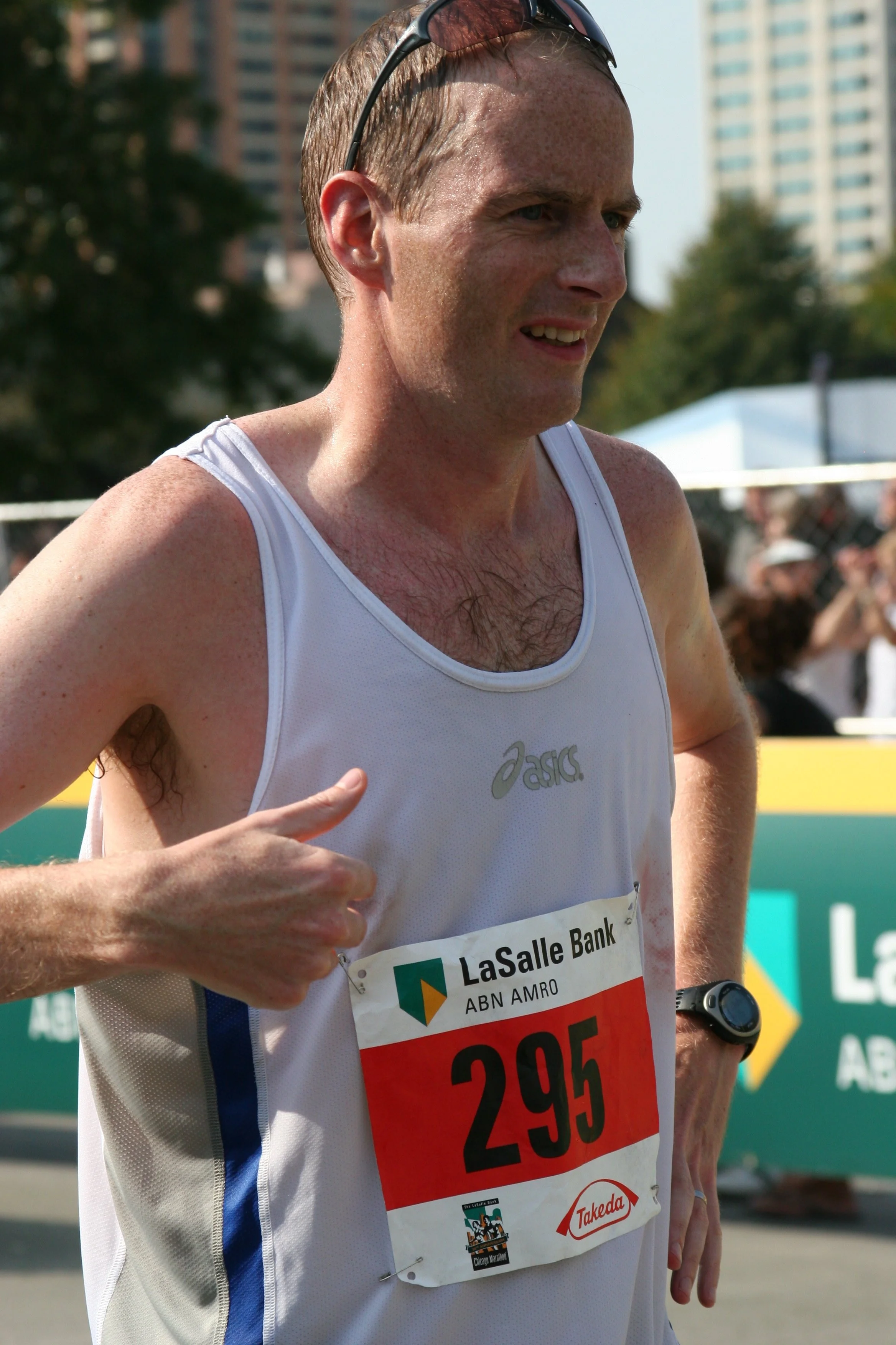 A male marathon runner wearing a white tank top with a race bib, sunglasses on his head, and a sports watch, running outdoors during a race with a cityscape and spectators in the background.