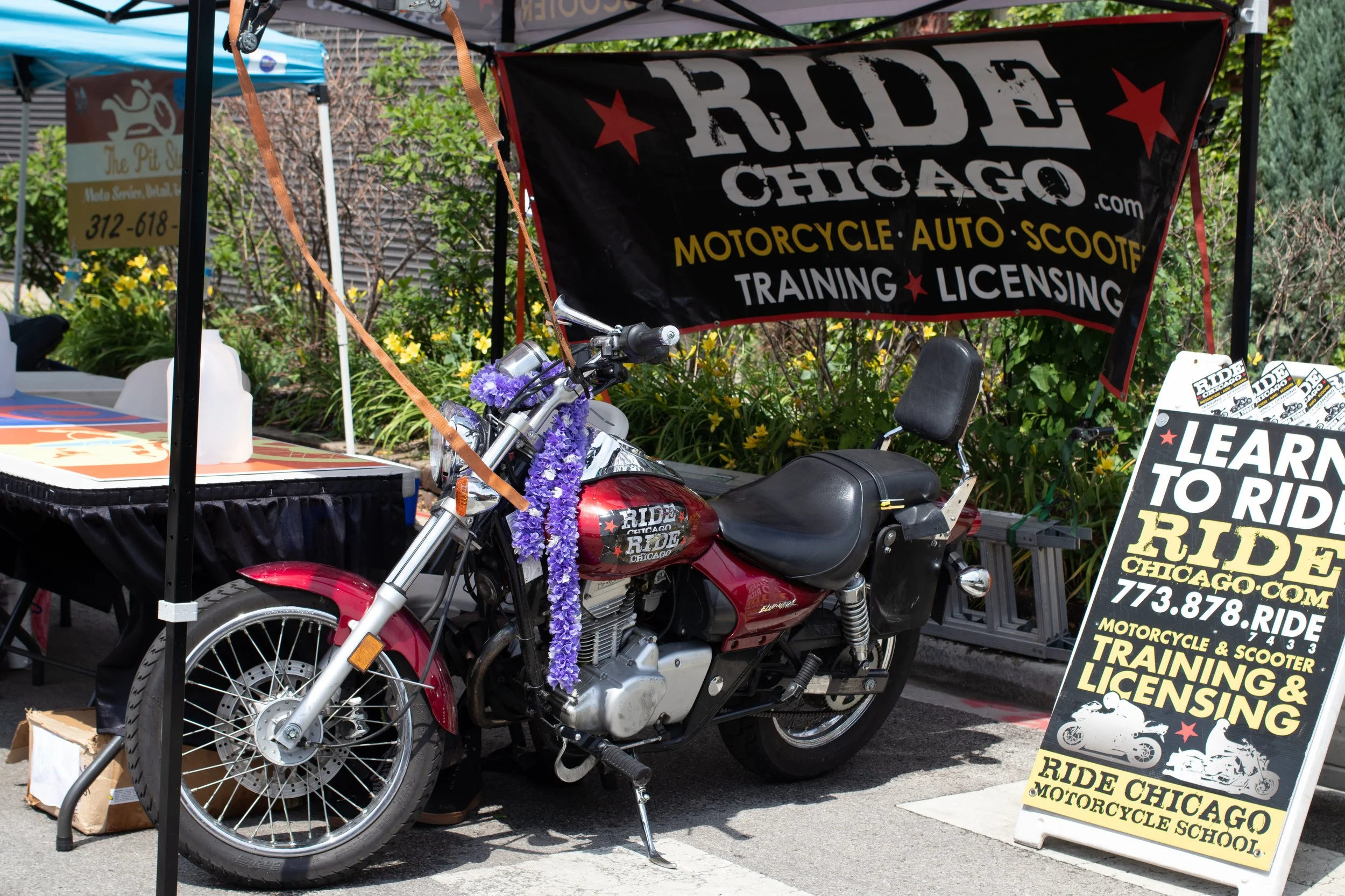 A red motorcycle parked under a tent at Ride Chicago Motorcycle School, with a signboard advertising riding lessons, training, and licensing services. The motorcycle is decorated with a purple flower lei.