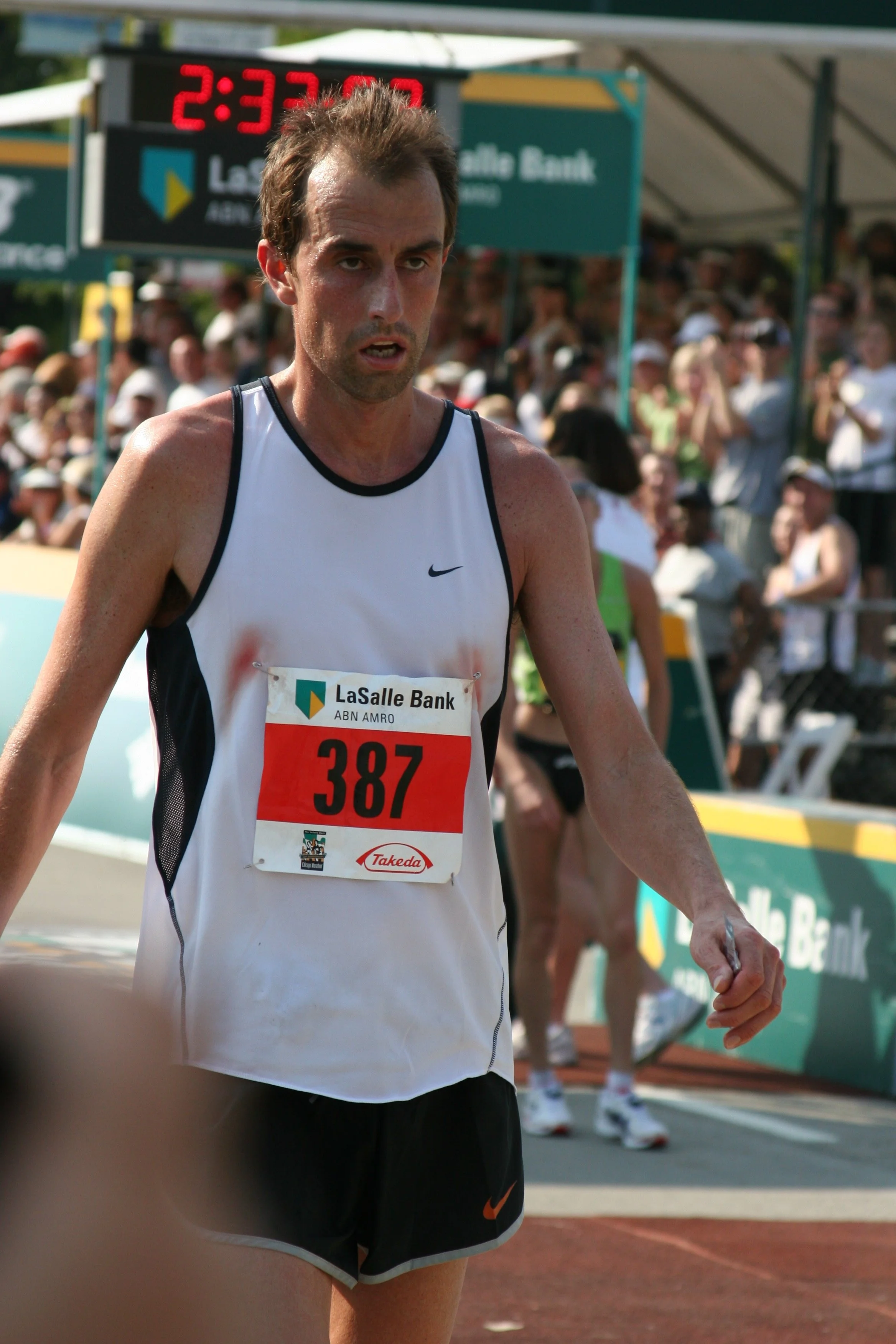 A male marathon runner is crossing the finish line, wearing a race bib numbered 387 with the logo for LaSalle Bank, with a crowd of spectators in the background.