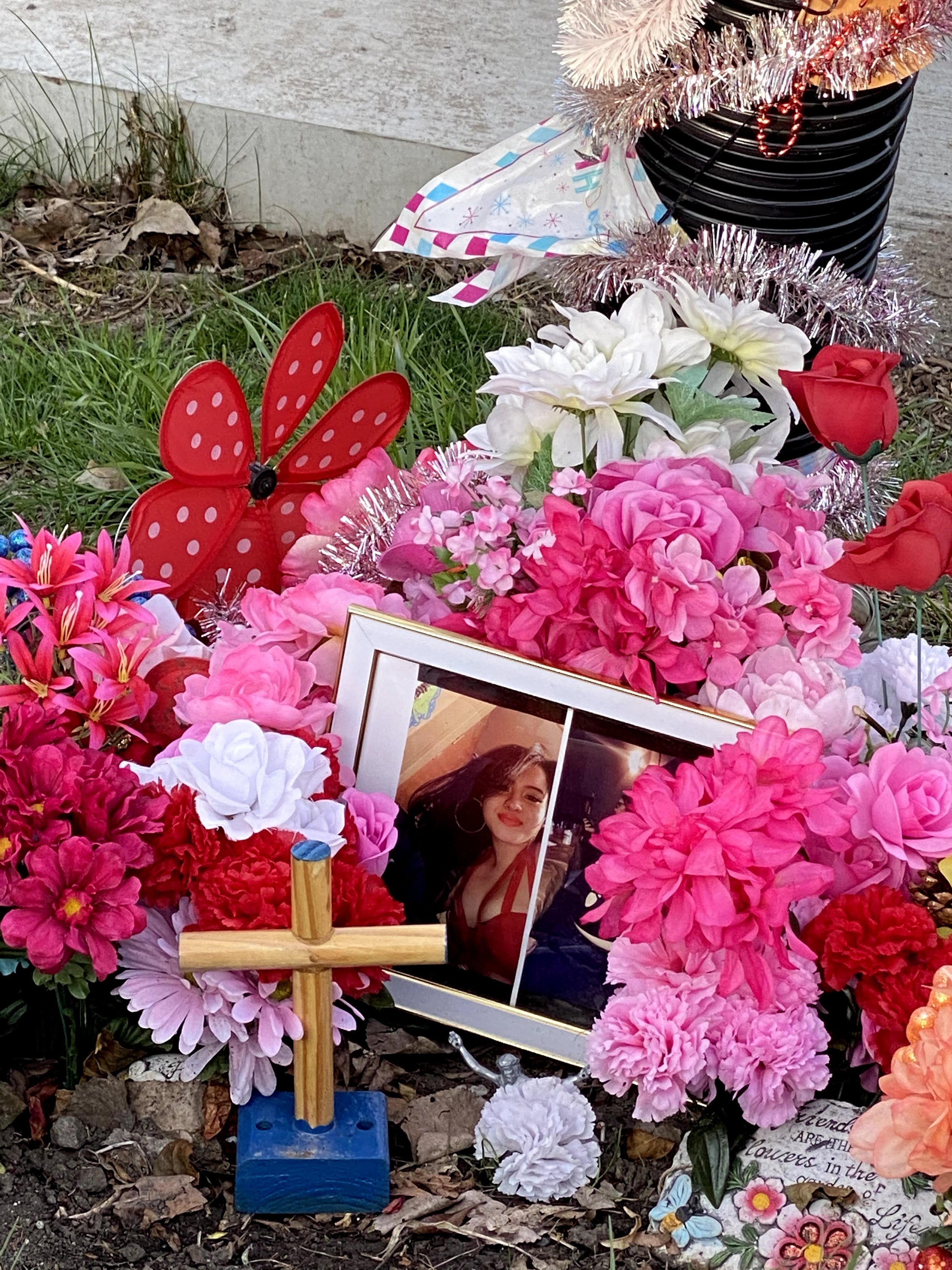A small memorial display with artificial pink, white, and red flowers, a framed photograph of a woman with dark hair, and a red butterfly decoration surrounded by greenery and fallen leaves.