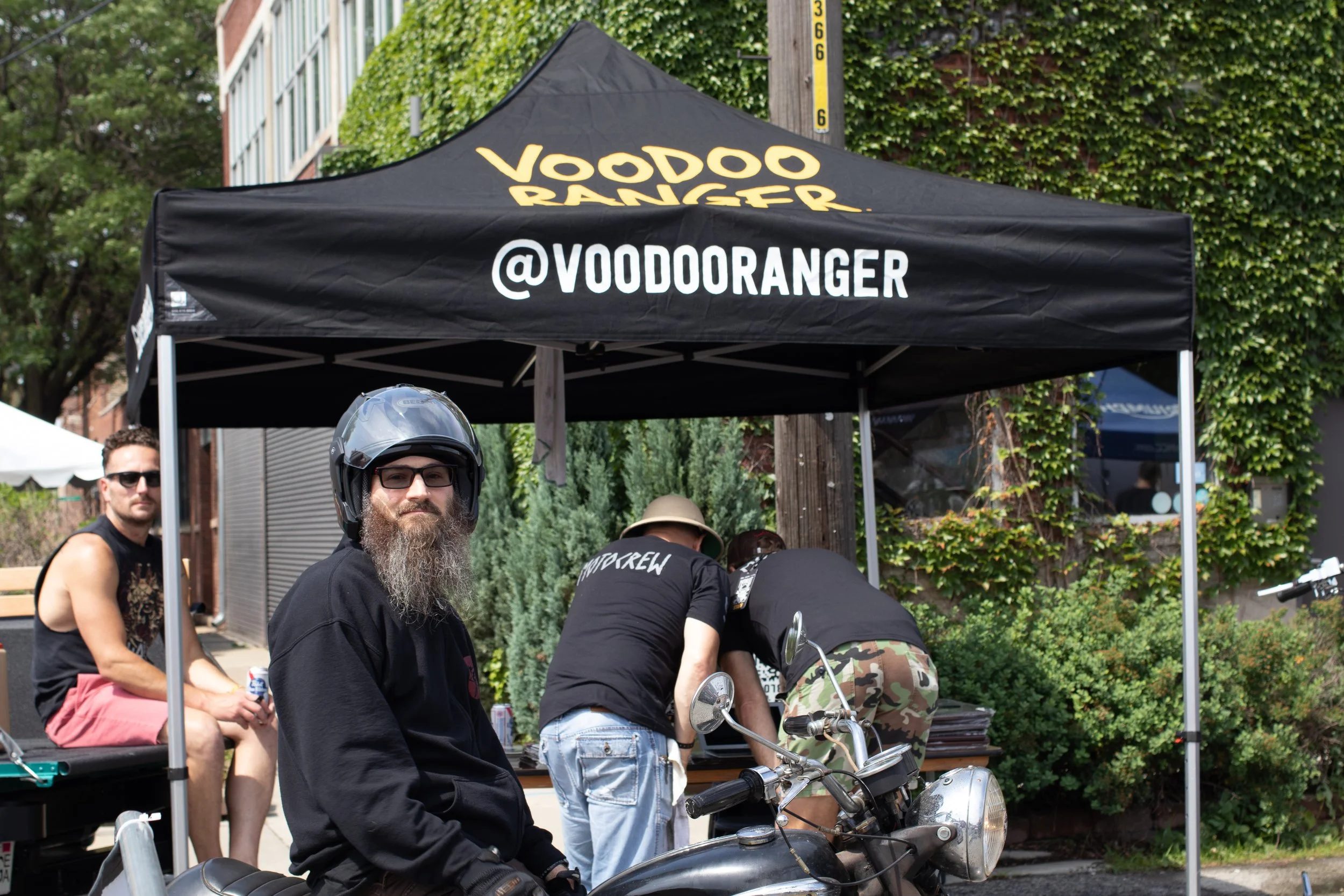 A group of people gathered at an outdoor event under a black canopy tent that reads 'Voodoo Ranger' and '@VOODOORANGER'. One man in the foreground, wearing glasses, a black helmet, and a black shirt, is sitting on a motorcycle. Other individuals are 