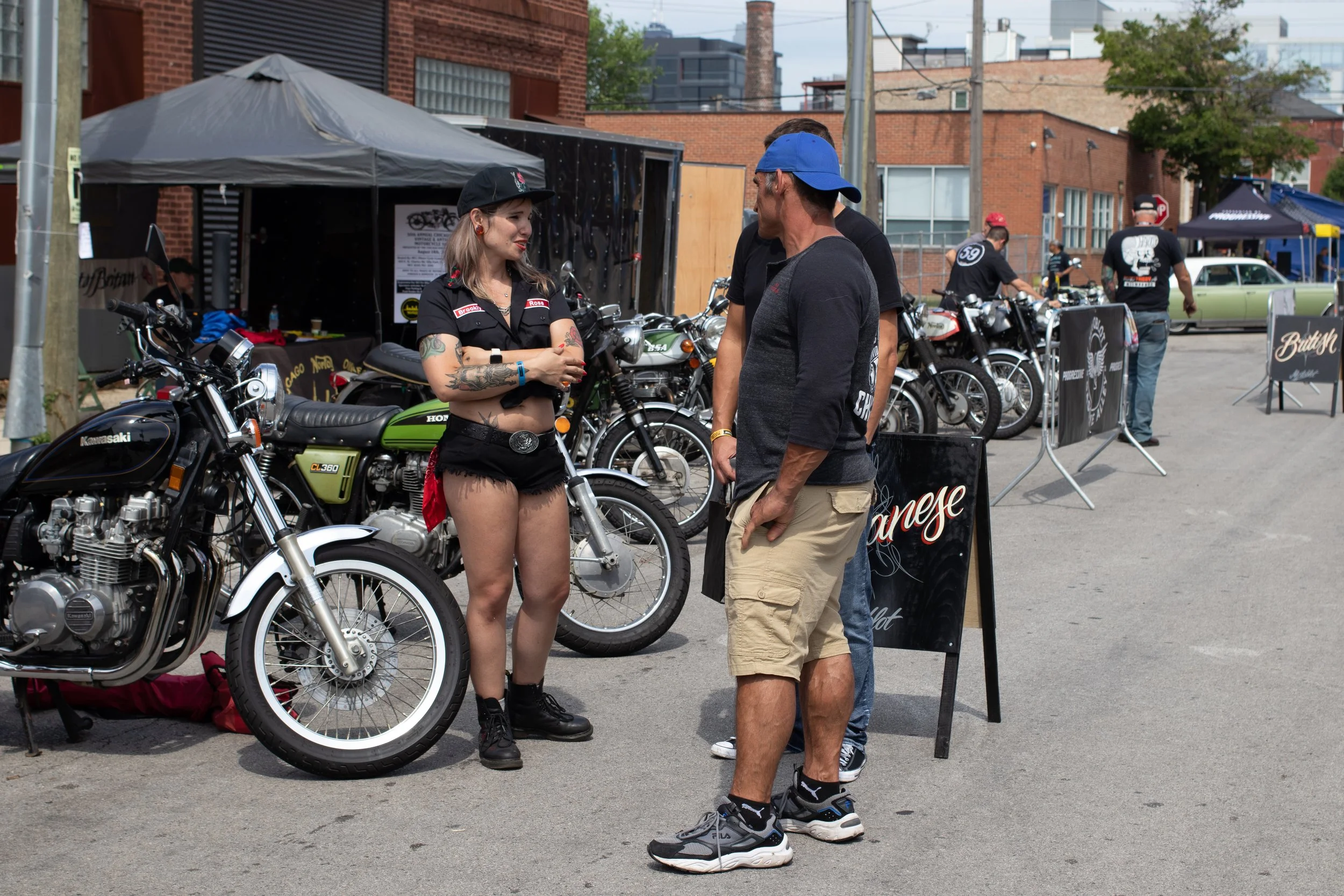 A woman with tattoos and dark hair wearing a black cap and black outfit talking to a man in a blue cap and beige shorts at a motorcycle event. Multiple motorcycles are displayed, and there are tents and people in the background.