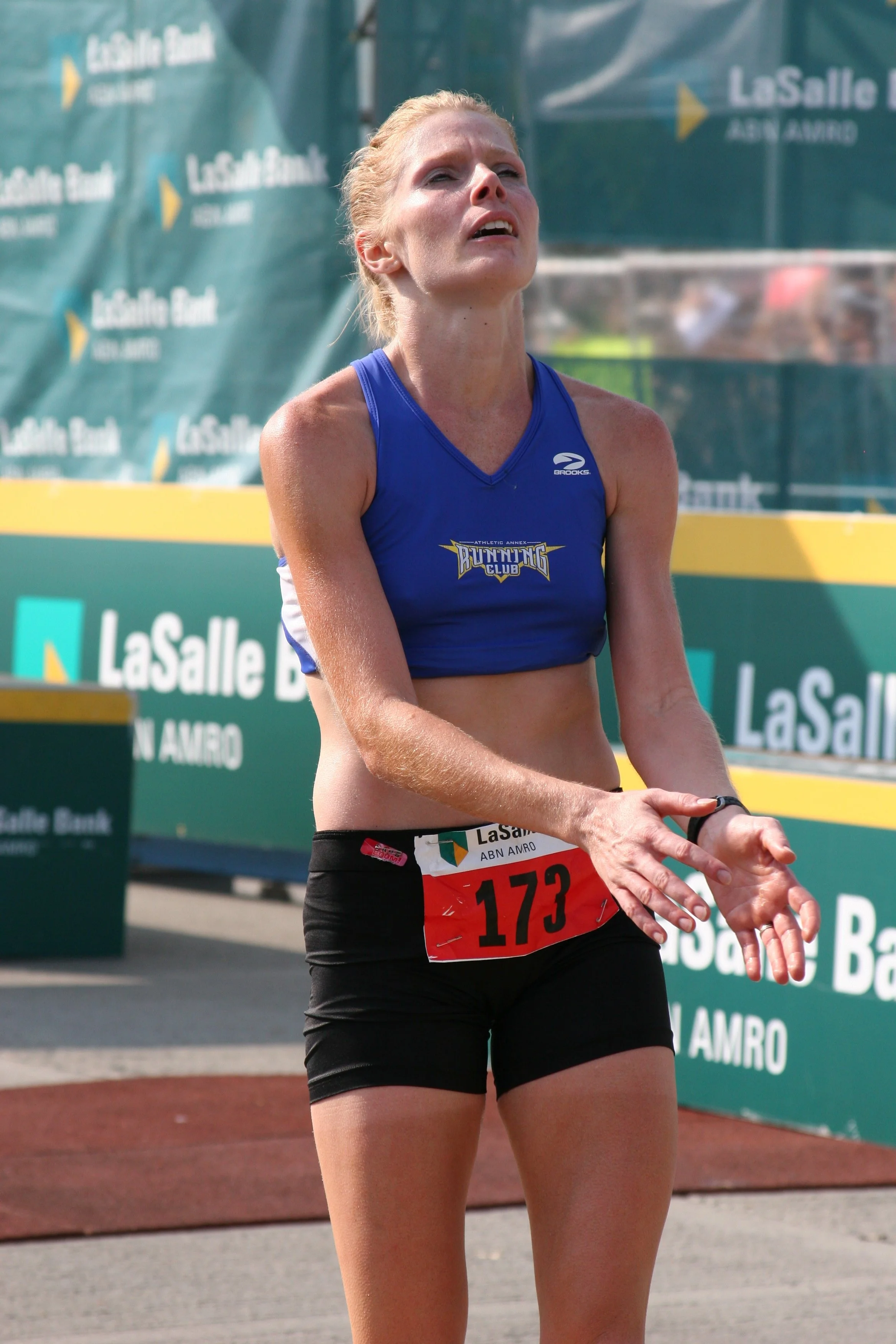 A female marathon runner with a race bib numbered 173, wearing a blue athletic top and black shorts, appears exhausted and is clapping while finishing the race, with the LaSalle Bank signage visible in the background.