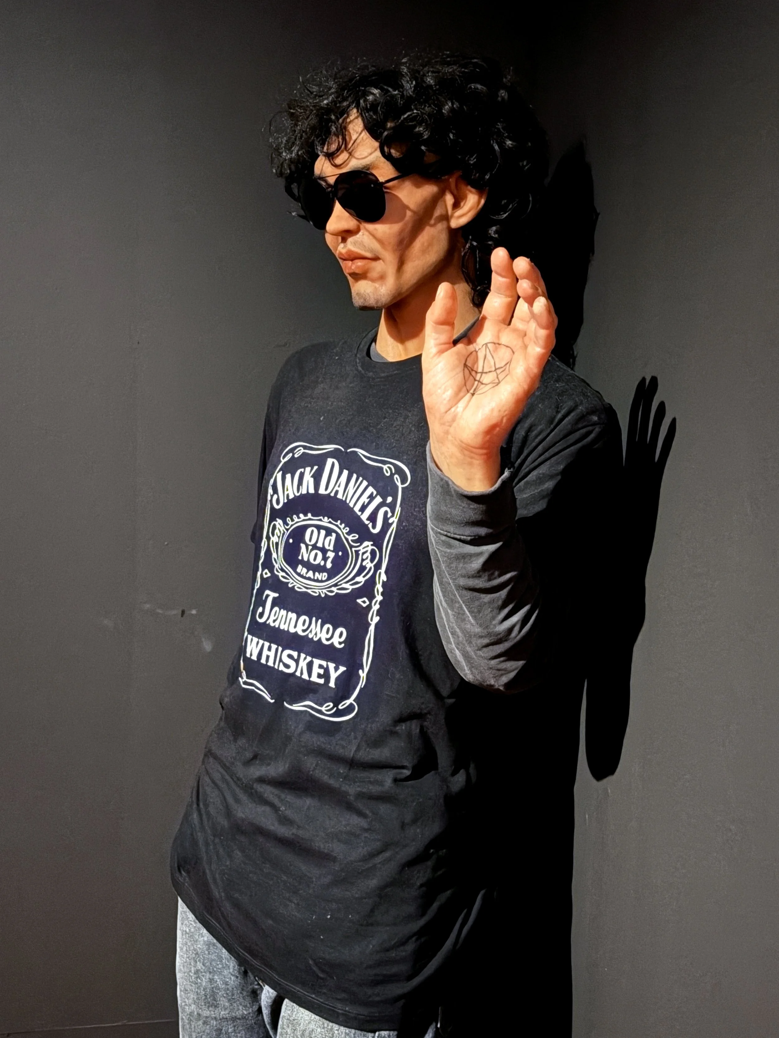 A man with curly black hair wearing sunglasses and a black Jack Daniel's t-shirt, raising his right hand with a diamond-shaped tattoo visible on his palm, standing against a plain dark background.
