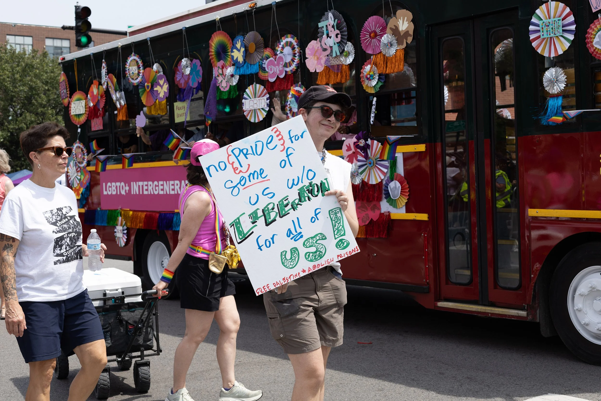 People participating in a Pride parade, standing in front of a decorated bus with rainbow-themed decorations and signs. One person holds a sign advocating for LGBTQ+ rights and against U.S. intervention in Palestine.