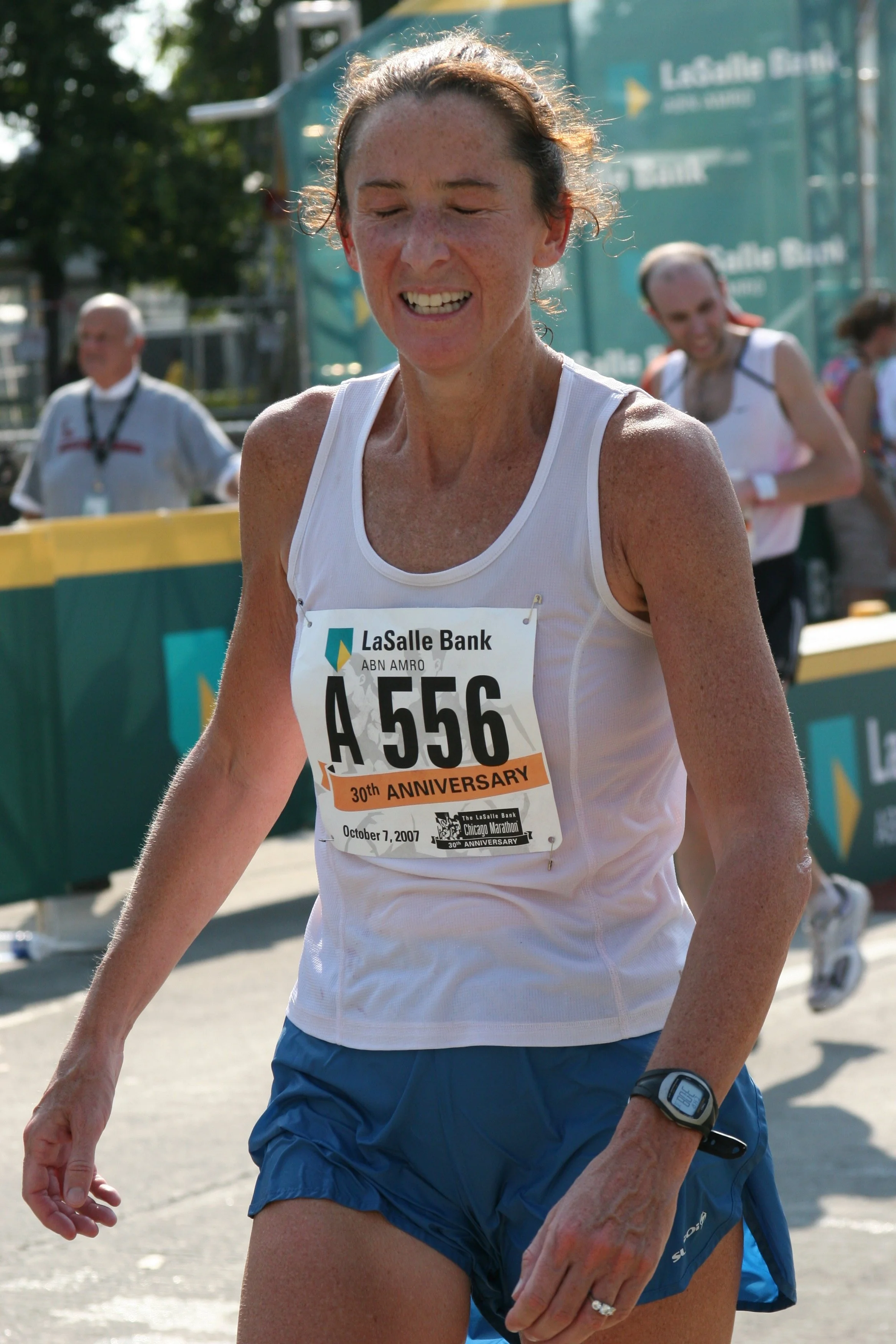 A woman with a race bib numbered A 556, smiling with her eyes closed, running in a race during daytime, wearing a white tank top, blue shorts, and a sports watch. The background includes other runners and a green tent.