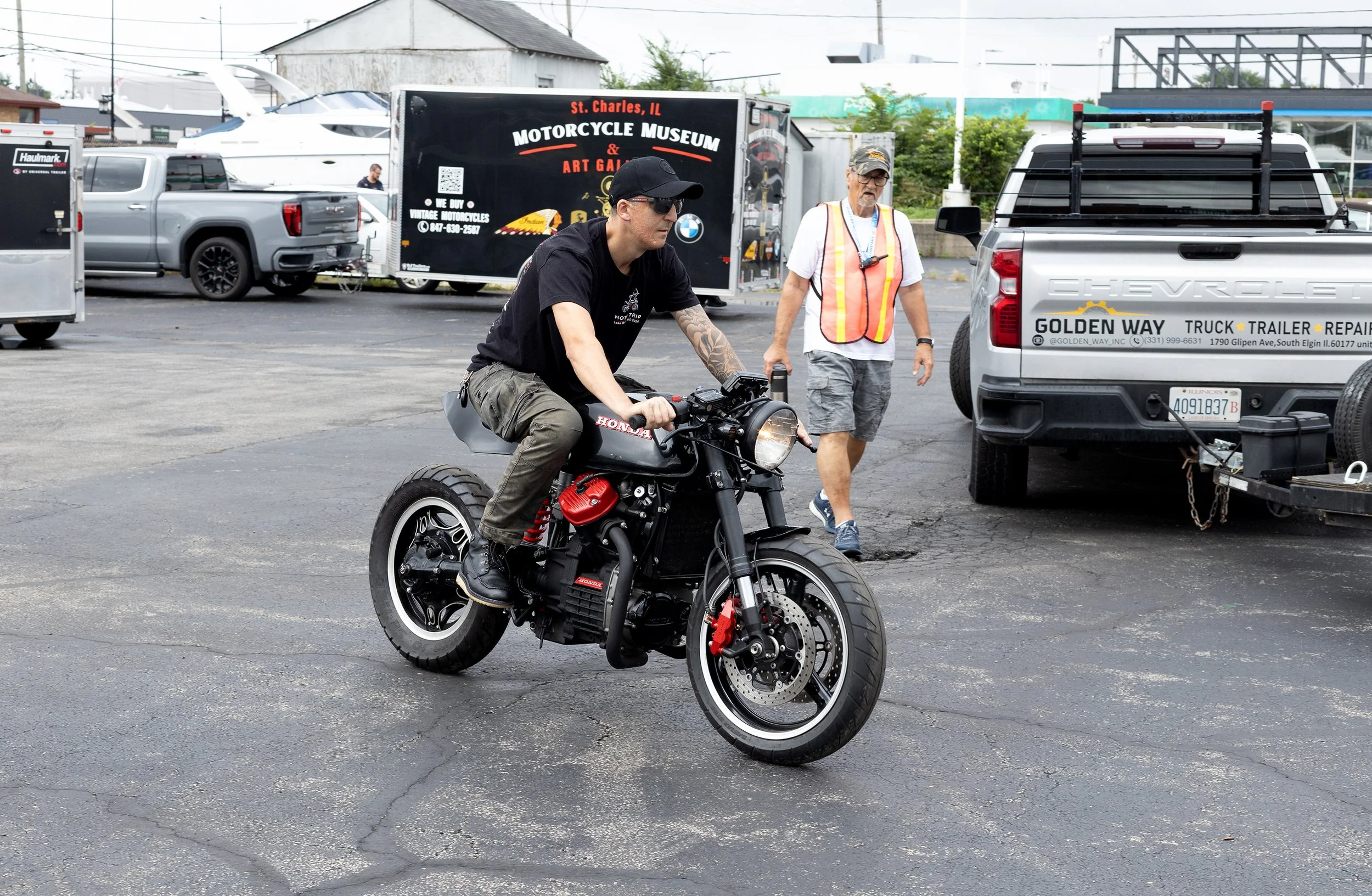A man wearing black riding a black Honda motorcycle with red accents, riding in a parking lot. Another man wearing a white shirt, gray shorts, sunglasses, and a high-visibility vest walks nearby, carrying a camera around his neck. The background show
