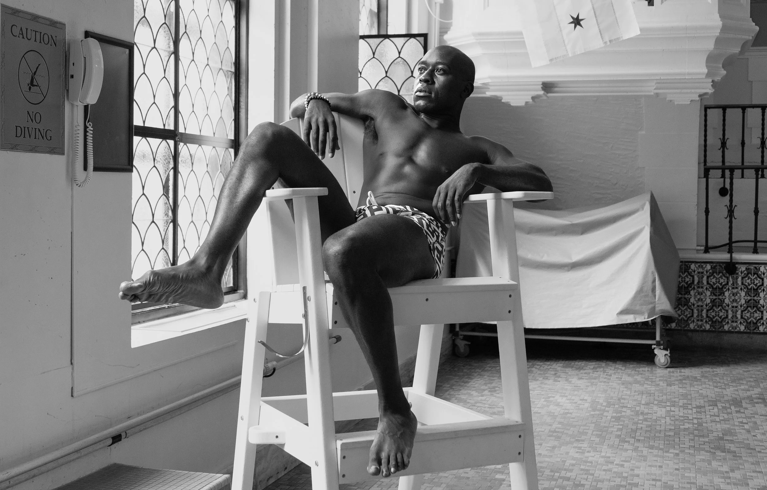 A black-and-white photo of a man sitting in a lifeguard chair inside a building. He is shirtless, wearing patterned swim trunks, and has a bracelet on his wrist. The man is posed with one arm resting on the side of the chair and the other hand on his