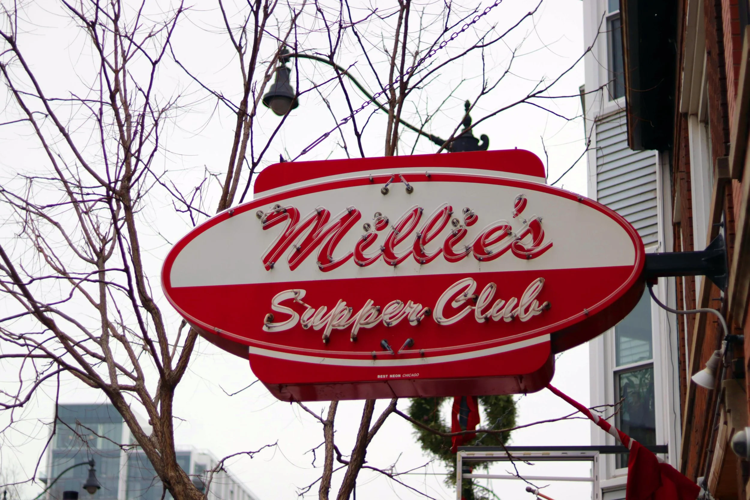 A red and white neon sign reading 'Miller'super Club' is hanging in front of a beige building, with leafless tree branches and a black lamp above it, on an overcast day.