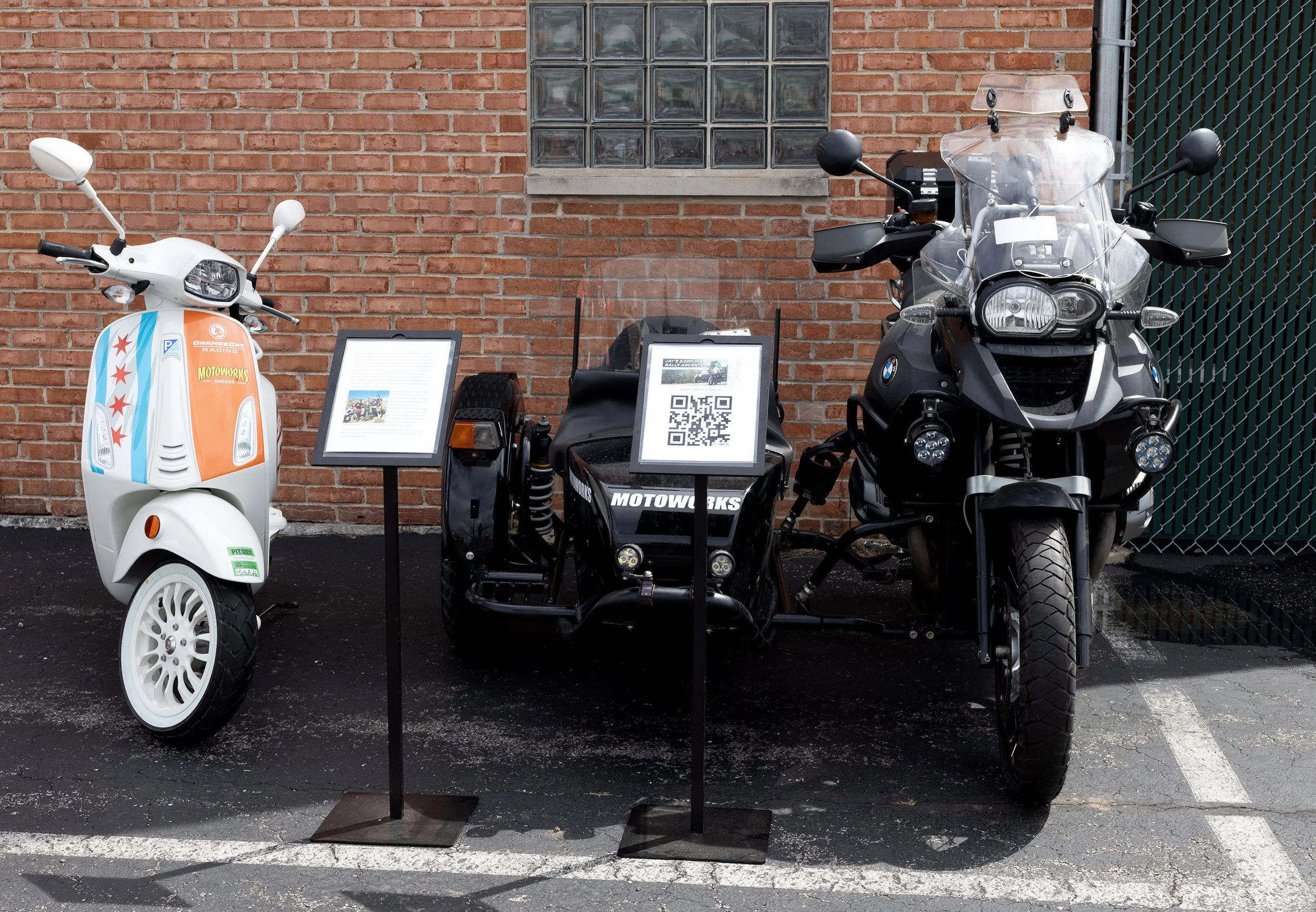 Display of two motorcycles and a scooter with informational signs in front of a brick wall.