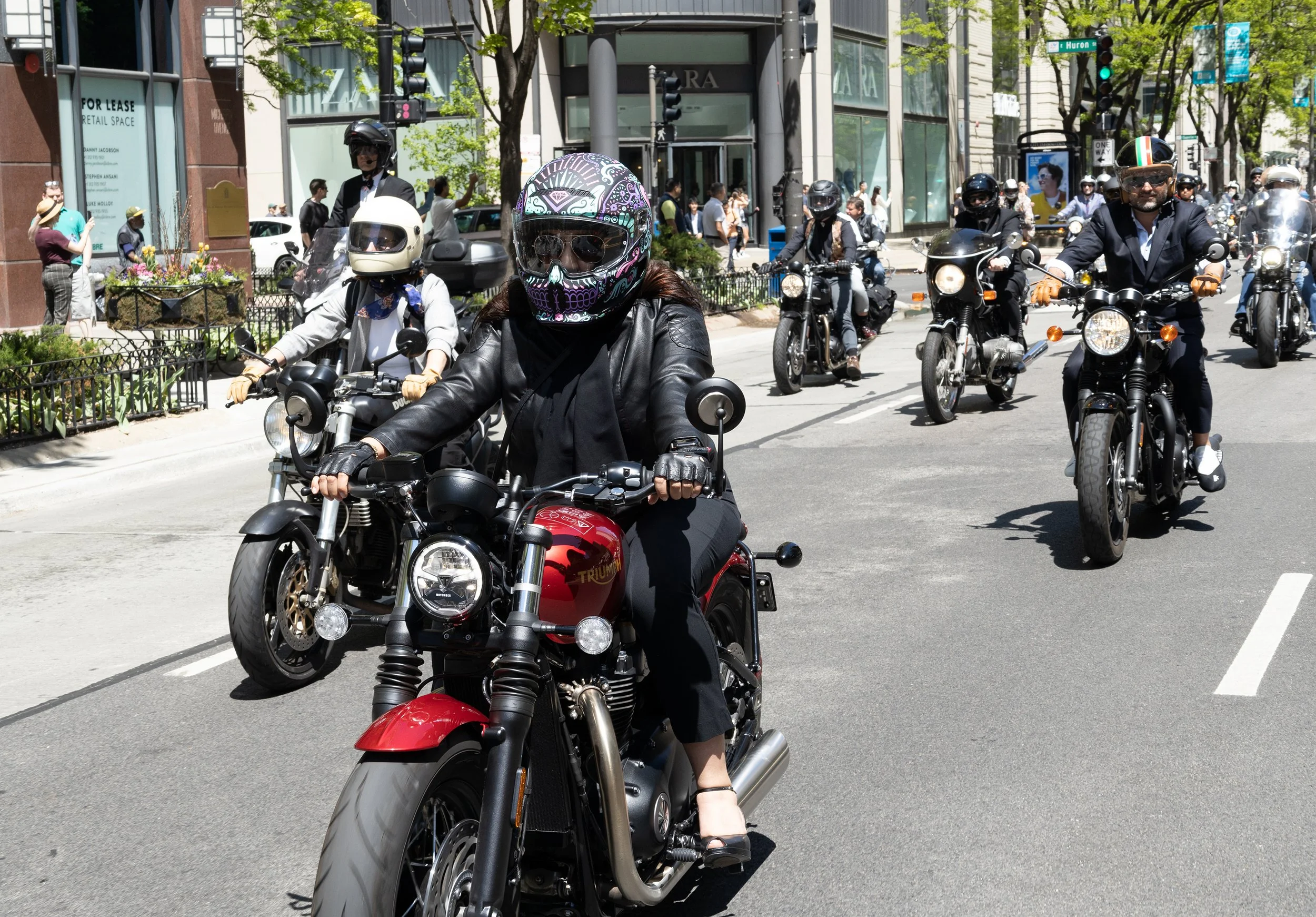 Multiple motorcyclists riding on a city street, with pedestrians walking on the sidewalk and buildings in the background.