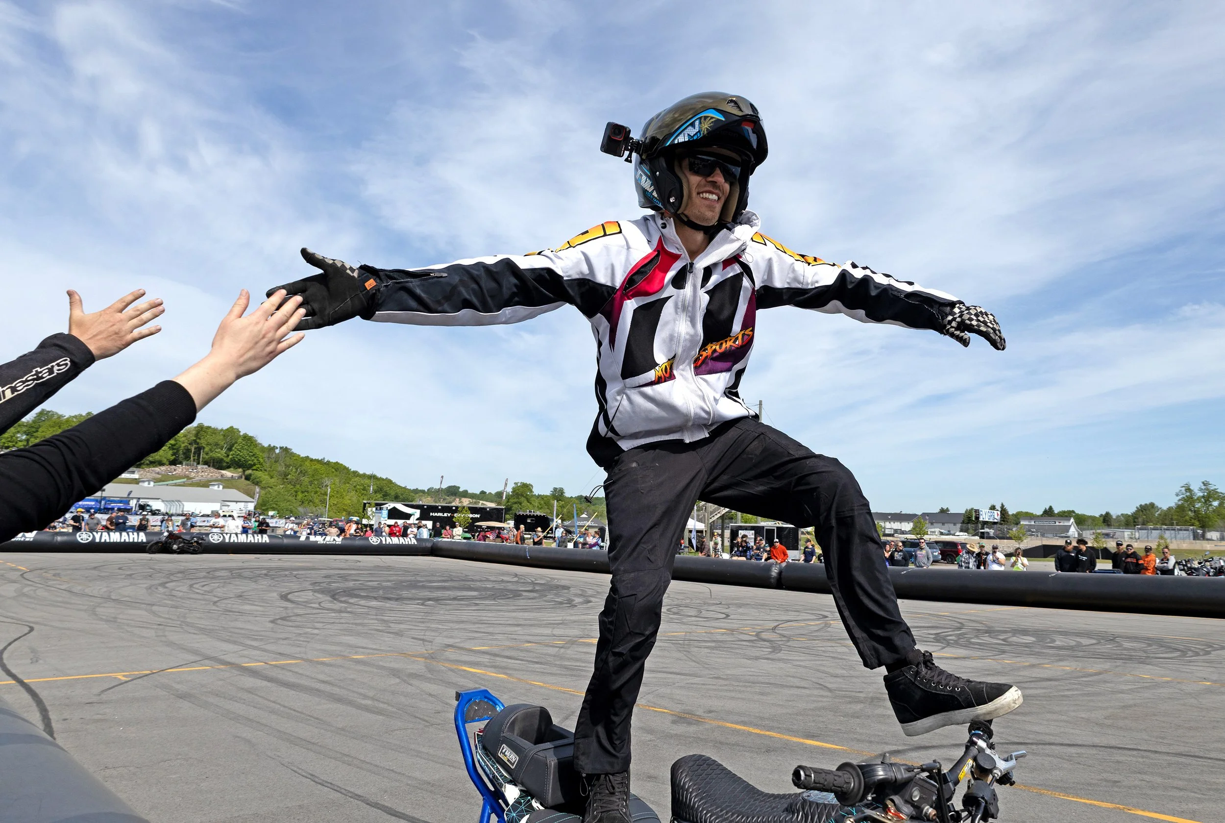 A man in motorcycle gear, including helmet and jacket, is standing on one leg with arms outstretched, in front of a crowd at a motorcycle event. A person with hands reaching out to him is visible, and a motorcycle is on the ground nearby. The sky is 