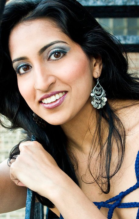 A woman with long black hair wearing large silver earrings and blue eyeshadow, smiling and looking at the camera.