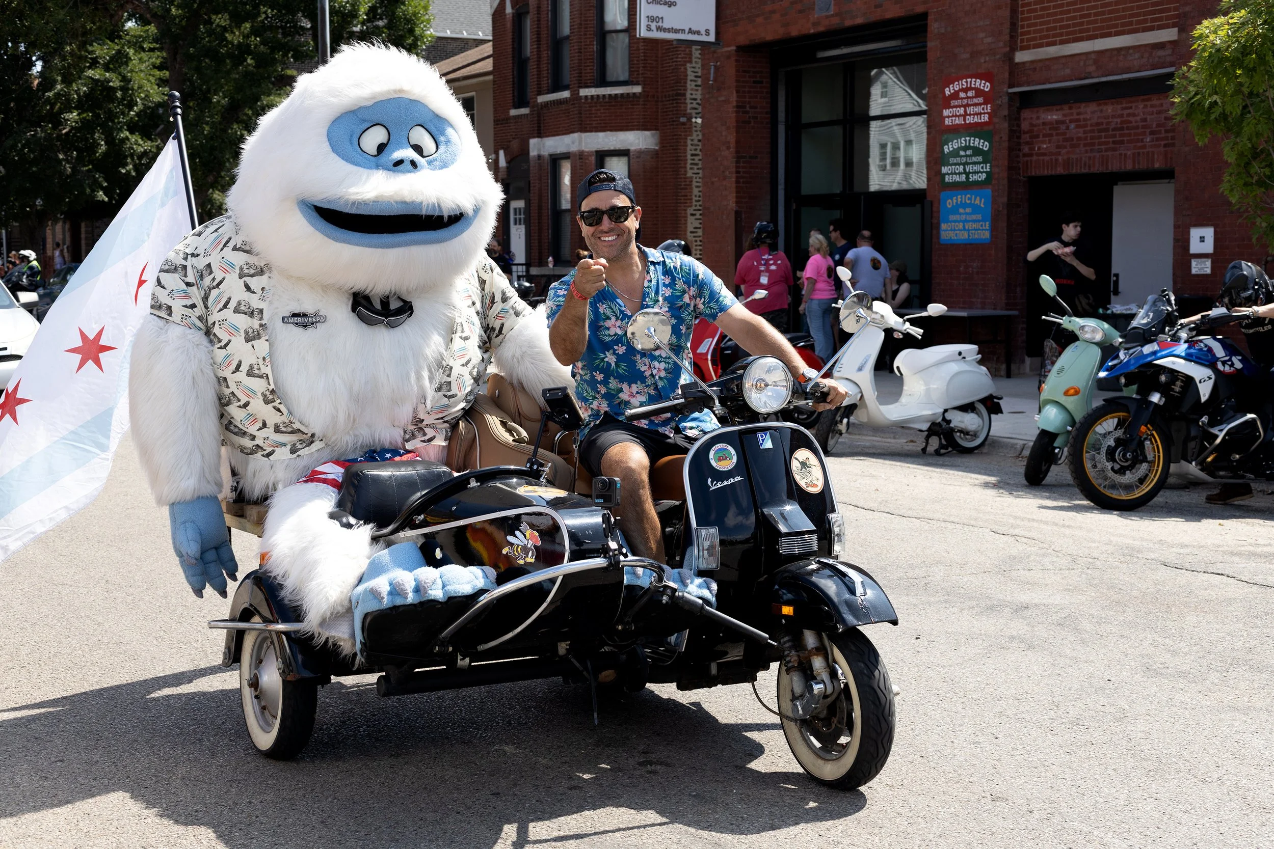 A person in a white gorilla mascot costume riding a motorized tricycle with a man in a blue Hawaiian shirt and sunglasses sitting on the bike. The mascot is holding a white flag with red stars. They are on a city street with parked scooters and motor