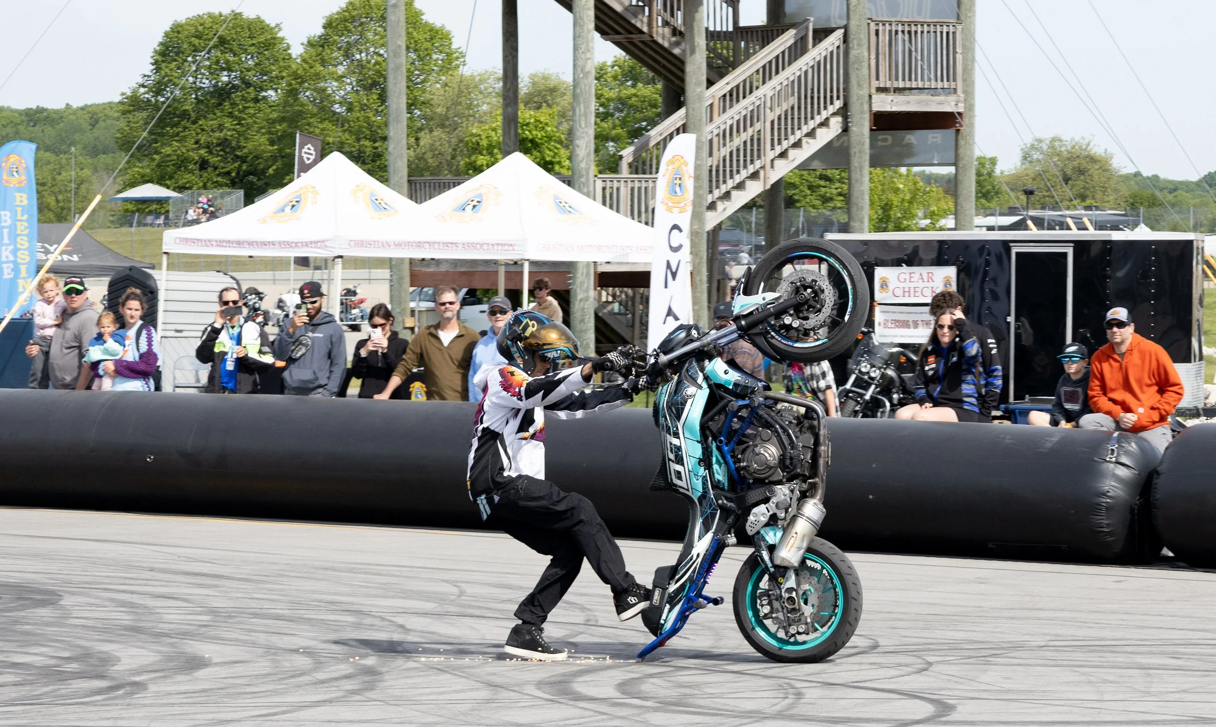 A motorcycle rider is performing a stunt on a motorcycle in an outdoor event, with spectators watching behind a safety barrier and tents in the background.