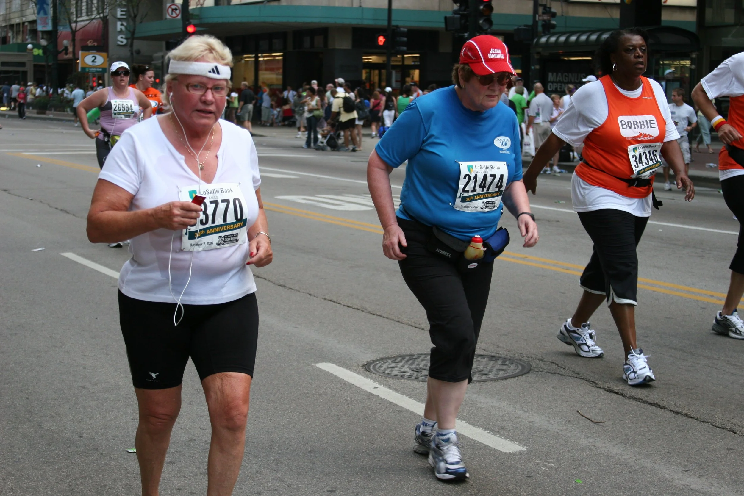 Three women running in a marathon, with a crowd and city buildings in the background. The women are wearing race bibs, athletic clothing, and accessories such as hats and headphones.