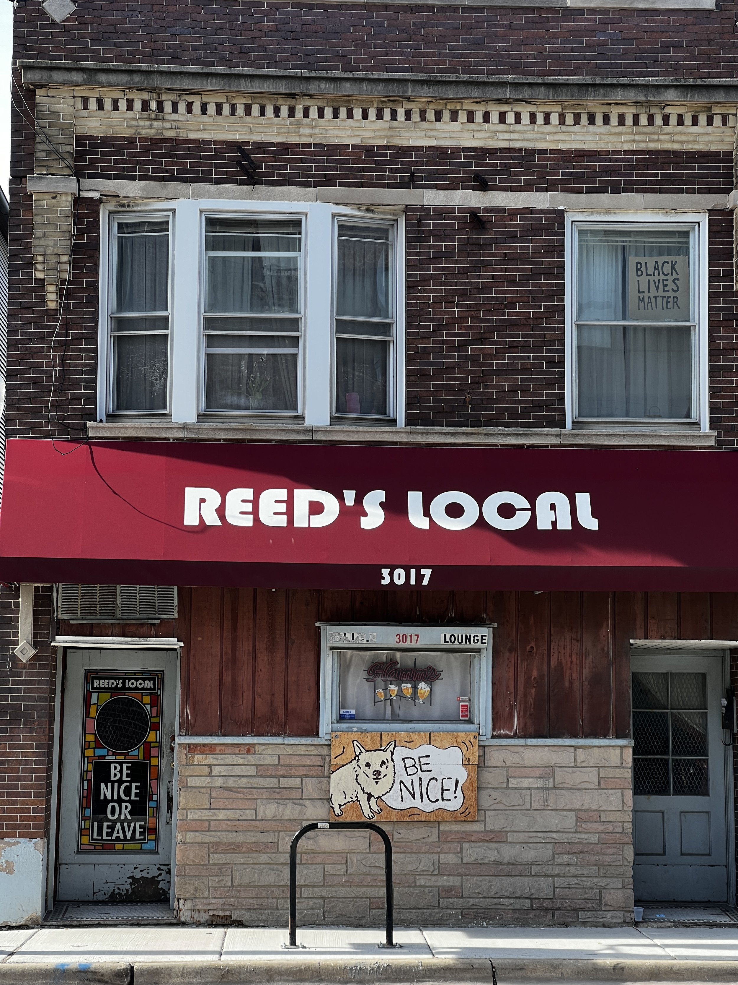 The image shows a brick storefront with a red awning that reads 'REED'S LOCAL'. There are two windows on the second floor, with one displaying a sign that reads 'BLACK LIVES MATTER'. Below, there is a door with a sticker that reads 'REED'S LOCAL' and