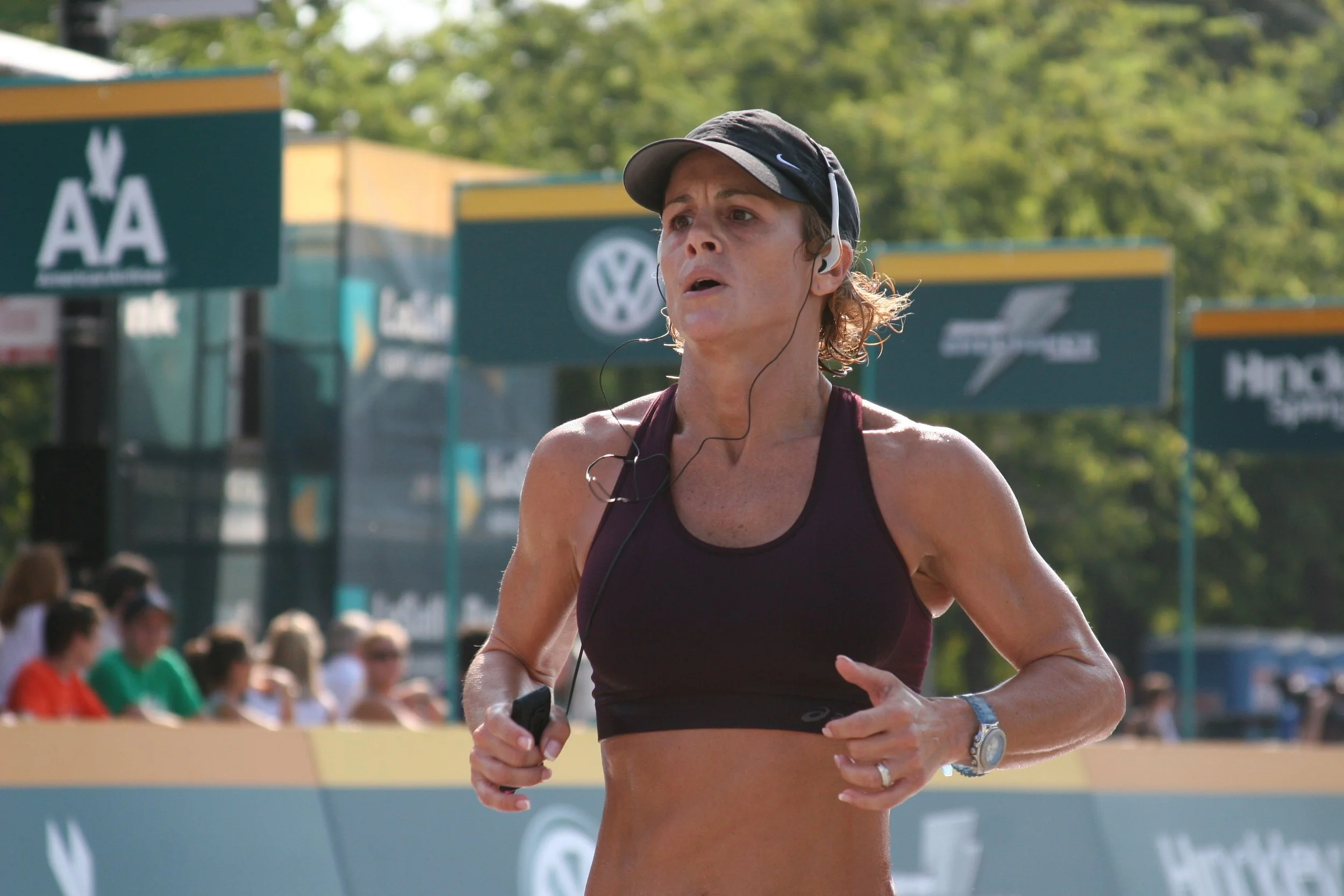 A woman running outdoors during a race or marathon, wearing a black cap, a maroon sports bra, headphones, and a wristwatch, with an audience and sponsor banners in the background.