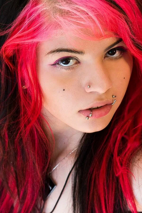 Close-up of a woman with bright pink and dark hair, wearing makeup and multiple facial piercings.