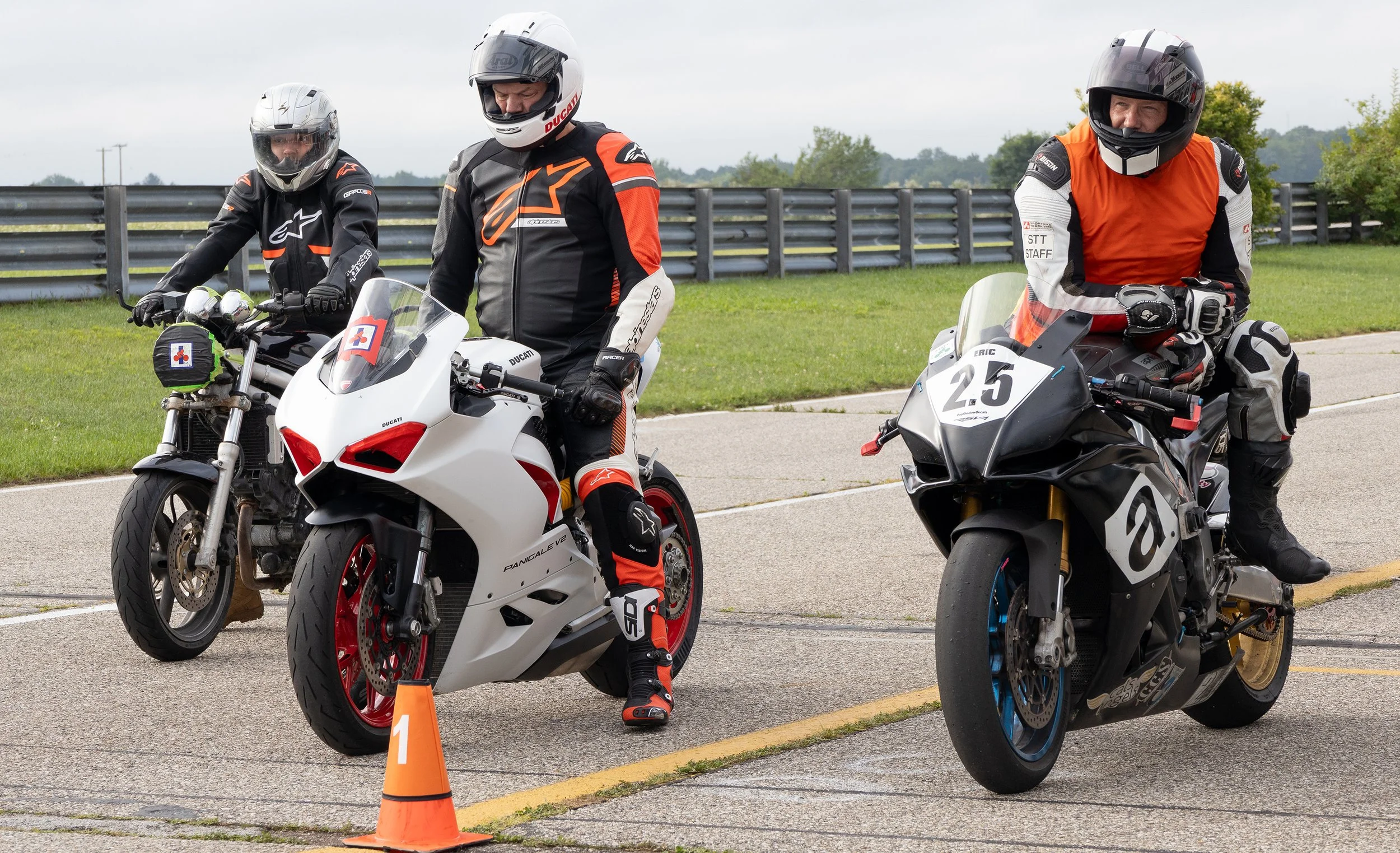 Three motorcyclists wearing protective gear and helmets, standing next to their bikes on a paved area with a grassy background and traffic cones.