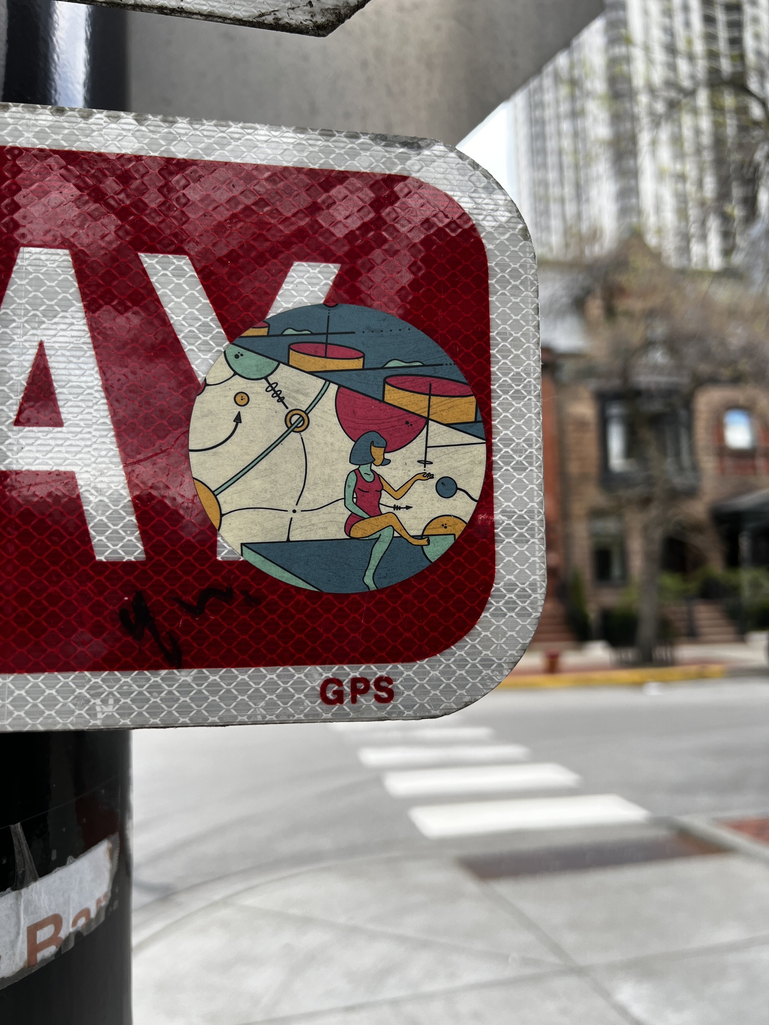 Close-up of a reflective traffic sign with a red and white diagonal crossing, showing a cartoon girl and a boat, in front of a pedestrian crosswalk and buildings.