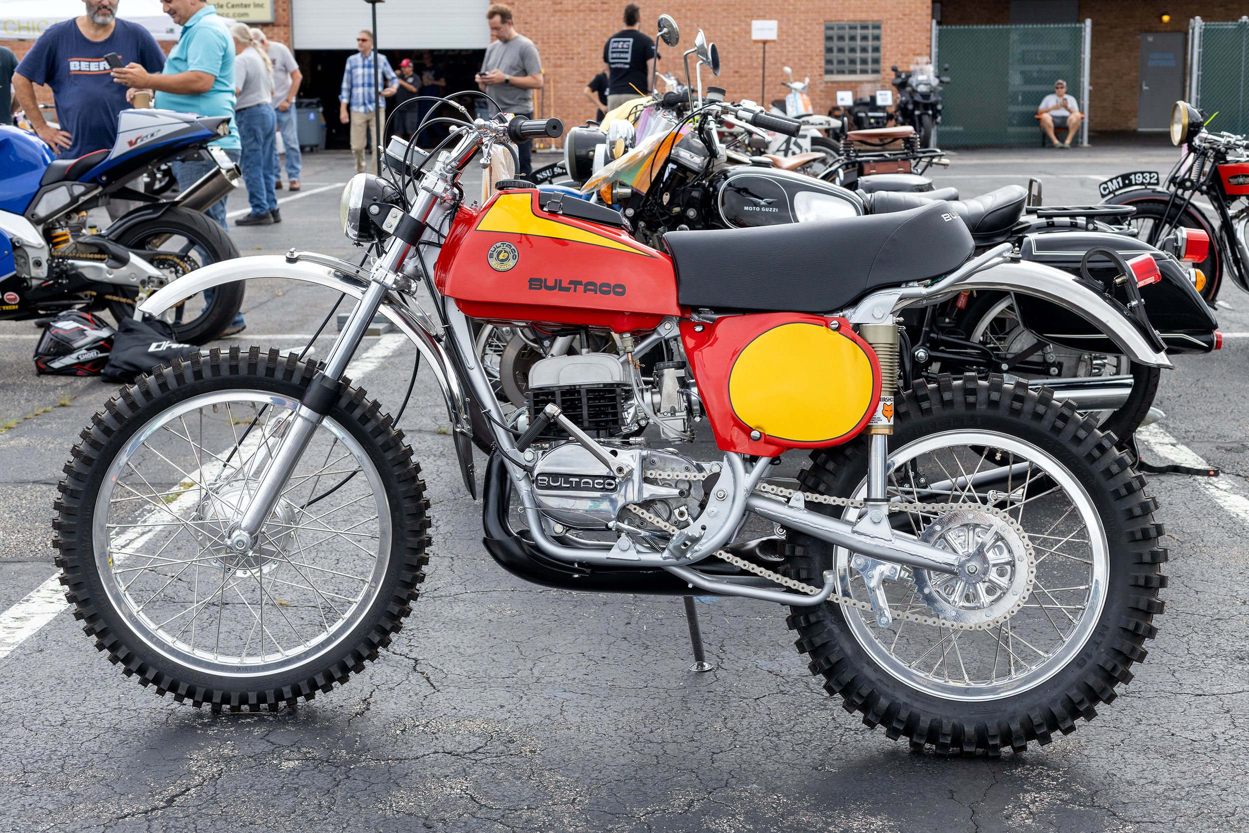 A vintage red and yellow Bultaco dirt bike with off-road tires, parked at an outdoor motorcycle event with people and other bikes in the background.