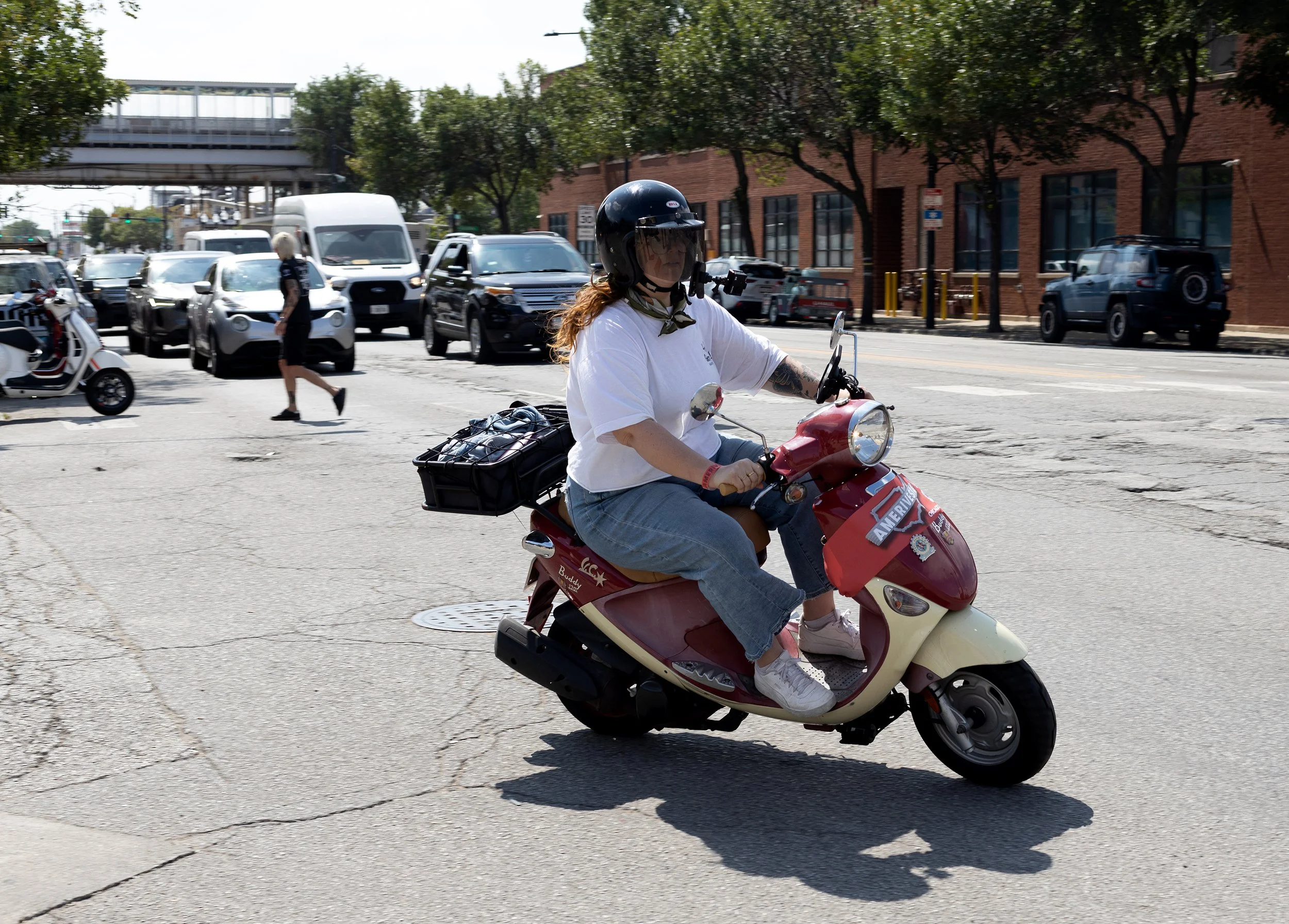 A woman wearing a safety helmet rides a red scooter on a city street, with cars and pedestrians in the background.
