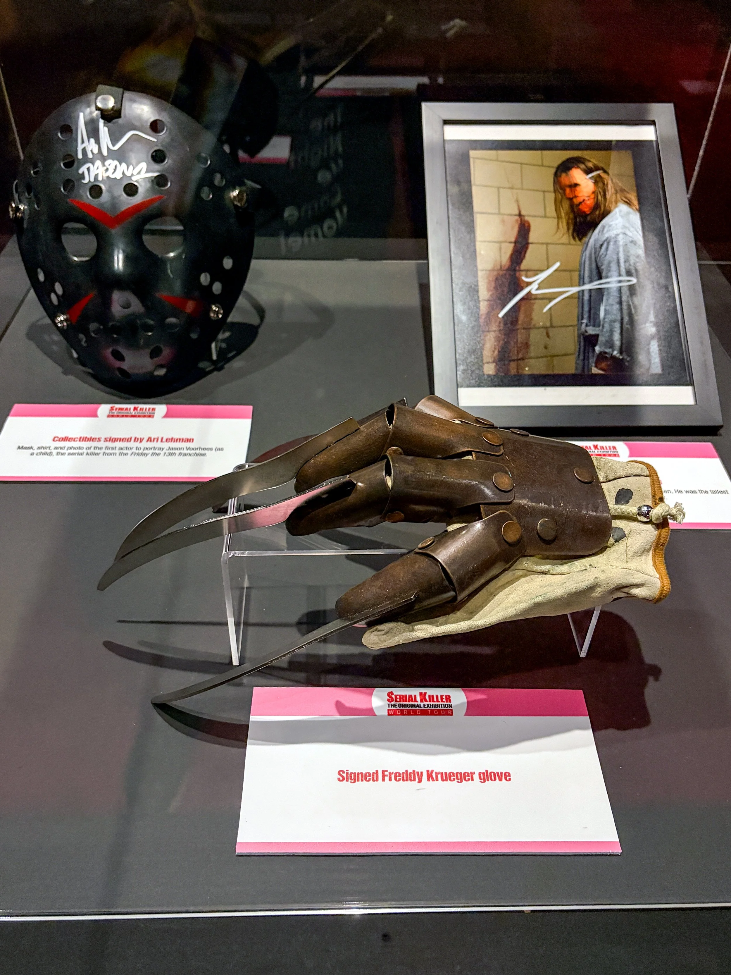 A display case containing a metal hockey mask with red markings, a signed Freddy Krueger glove with blades, and a framed photo of a person with long hair wearing a striped shirt. The display labels include titles such as "Serial Killer" and descripti