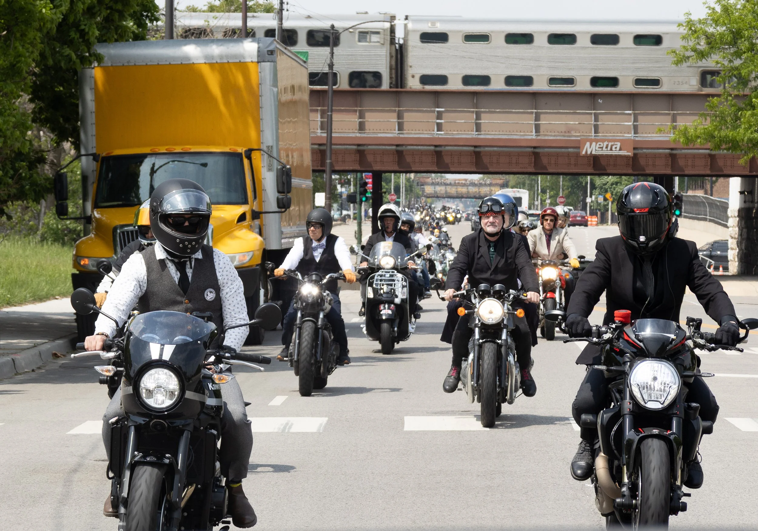 A group of motorcyclists riding together on a city street, with a yellow delivery truck and train passing overhead on an elevated railway.