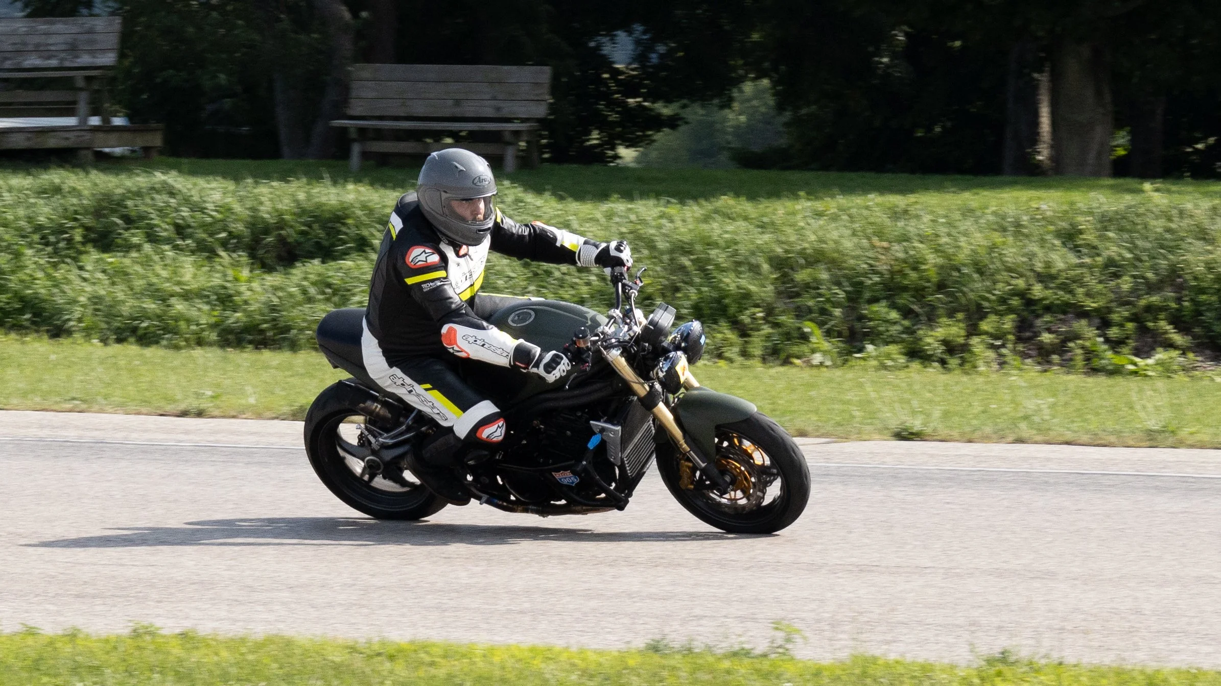 Motorcycle rider wearing a helmet and riding gear on a black motorcycle on a paved road with green grass and trees in the background.