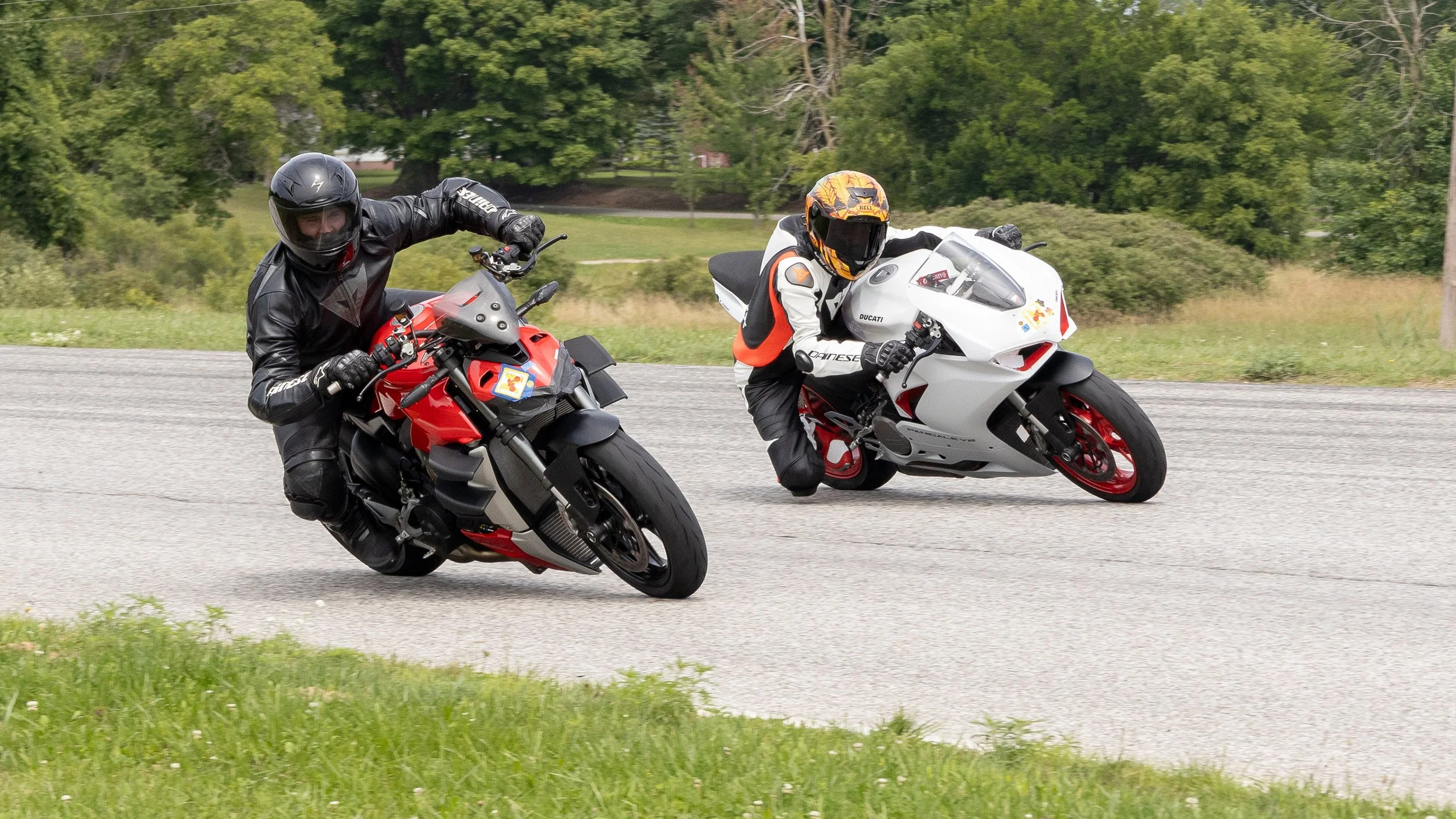Two motorcycle racers leaning into a turn on a paved road surrounded by greenery.