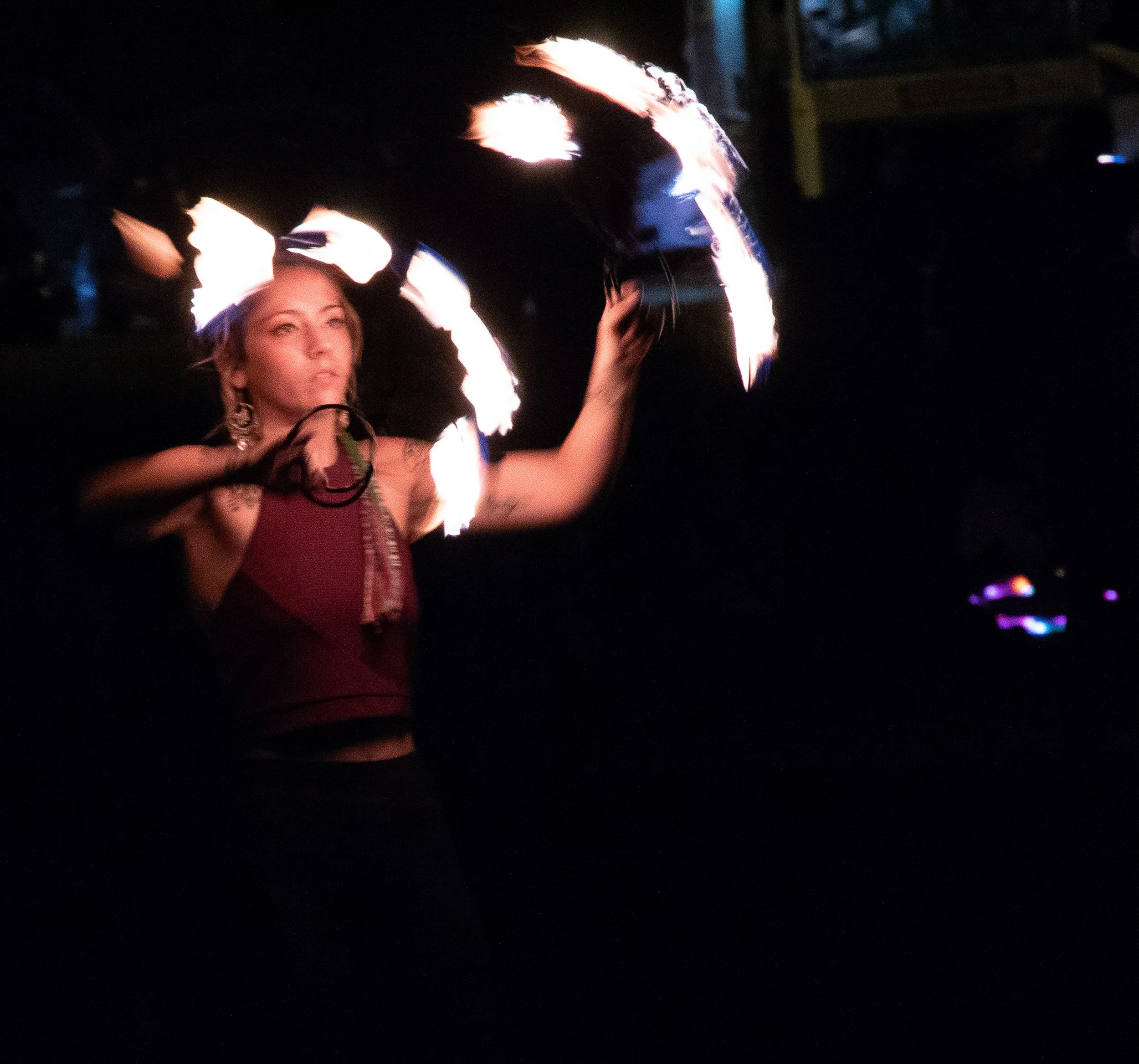 A woman with earrings and a patterned top holding a flamethrower with flames in a dark environment.
