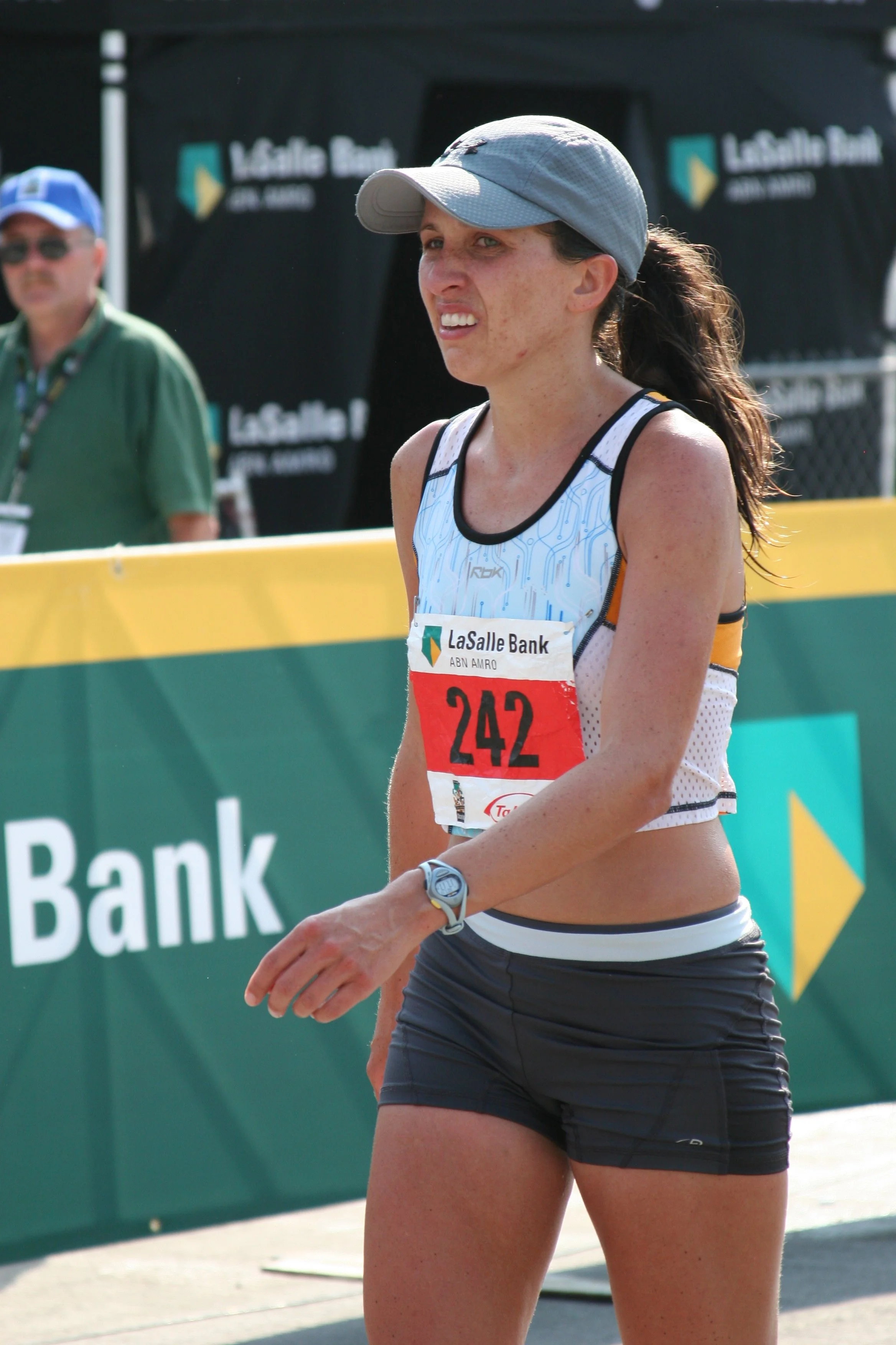A female athlete in running attire, wearing a gray cap and a race bib numbered 242, standing outdoors near a LaSalle Bank branded barrier, with a man and a black backdrop in the background.