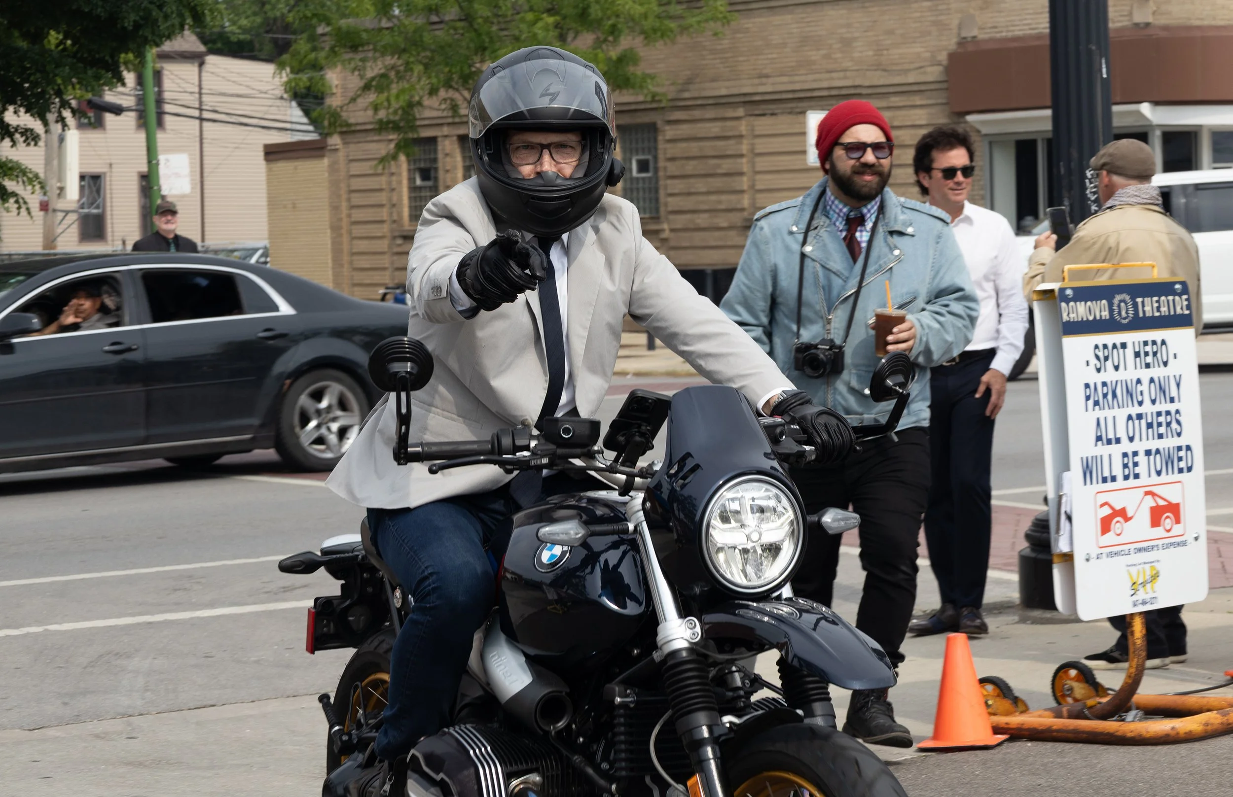 A man in a grey blazer and motorcycle helmet riding a BMW motorcycle stalls at an outdoor parking lot with other people, a car, and a sign indicating parking restrictions.