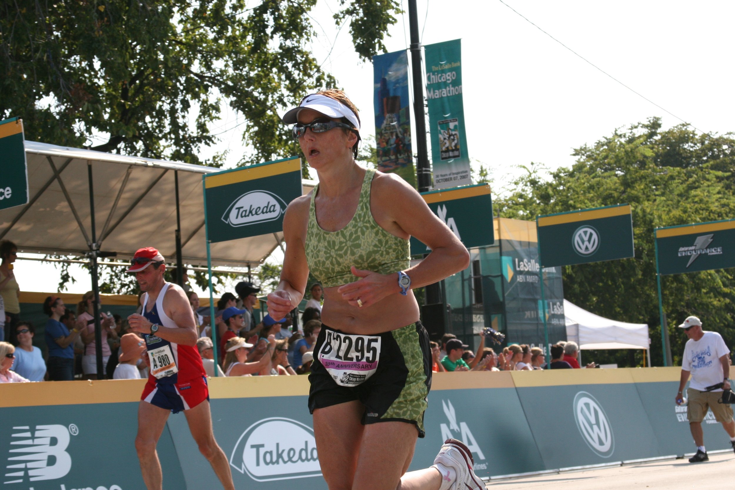 A woman running in a marathon, wearing a green athletic top, black shorts, sunglasses, a white cap, and a race bib numbered 11295, during daytime with trees and banners around the race area.