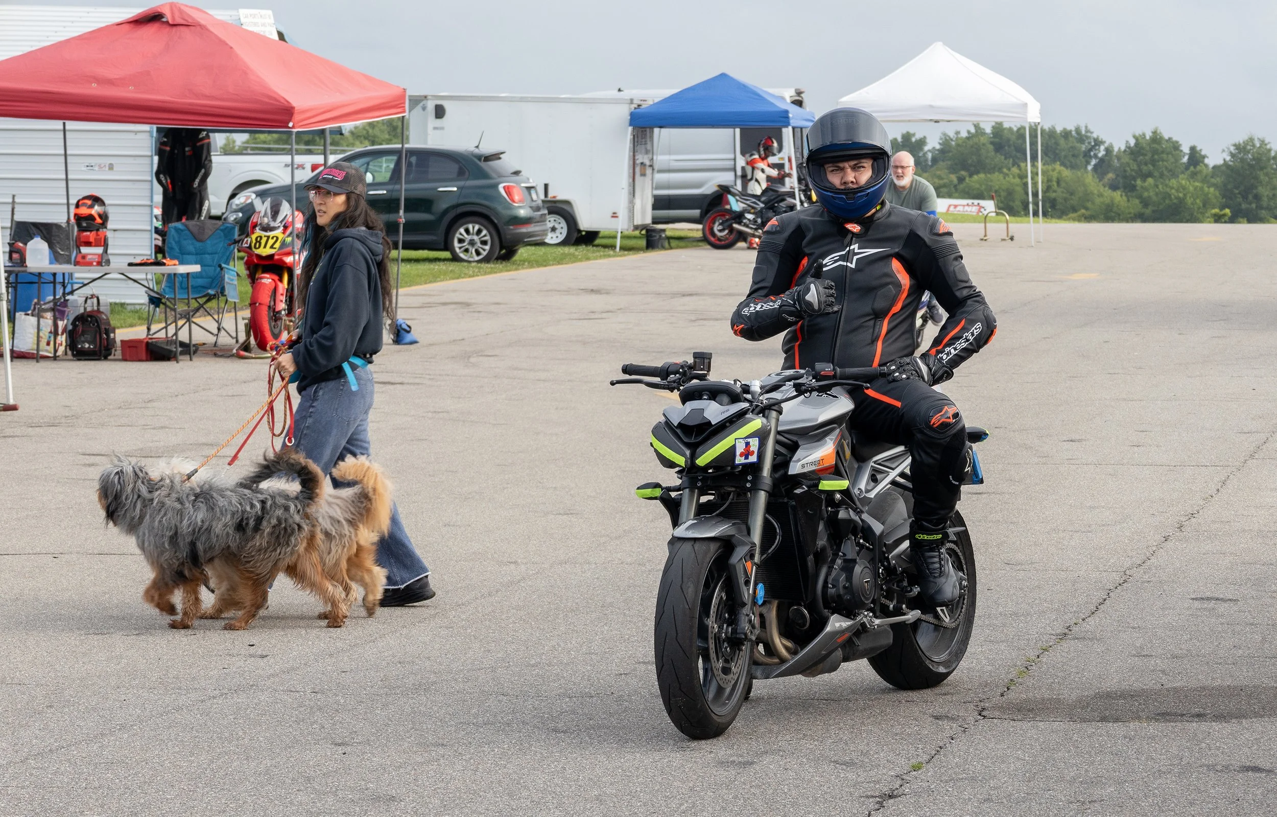 A man wearing motorcycle gear and helmet sitting on a black motorcycle with green accents in an outdoor parking lot. Behind him, a woman with glasses and a hat is walking a dog. There are canopies, vehicles, and motorcycles in the background.