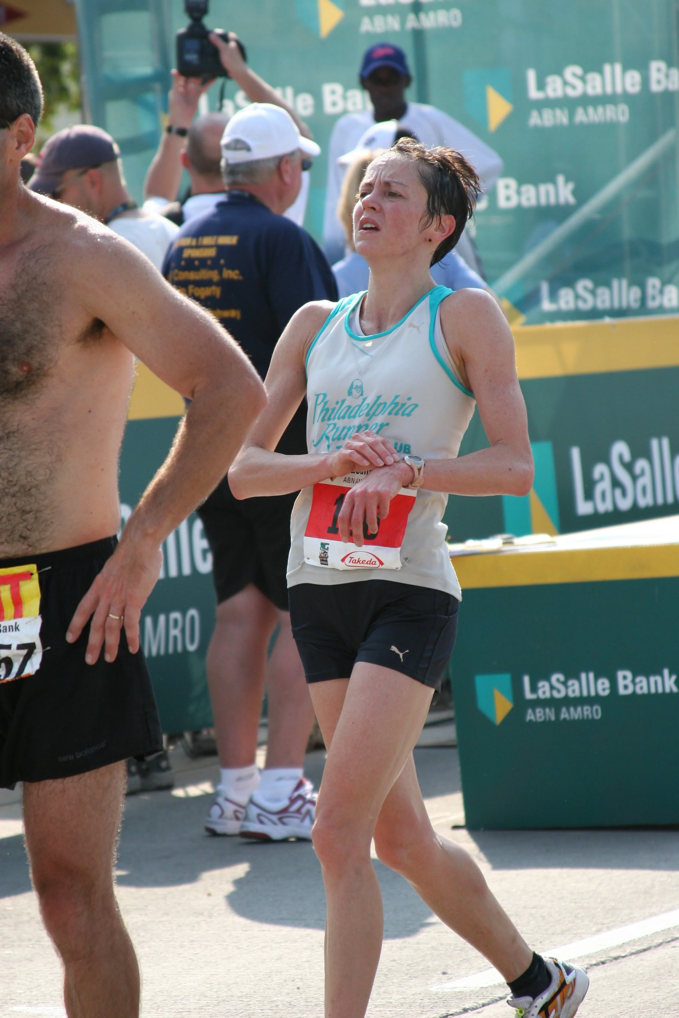 A woman in running gear, wearing a white shirt with blue accents, black shorts, and running shoes, participating in a marathon or race event. She appears to be catching her breath or cooling down. In the background, there are other runners, event sta