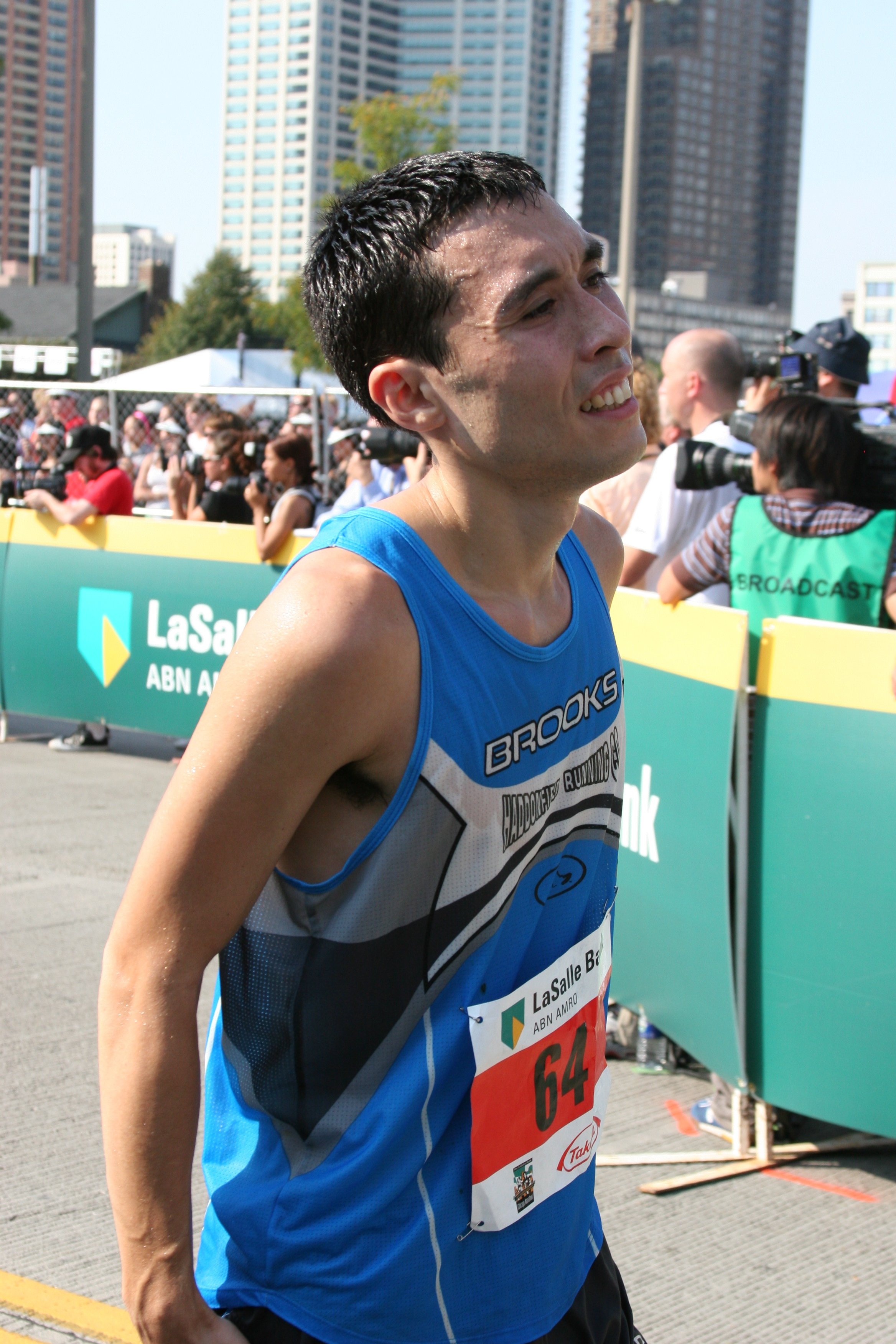Male marathon runner in blue tank top with race bib, sweating, during a race with city buildings, spectators, and cameras in background.