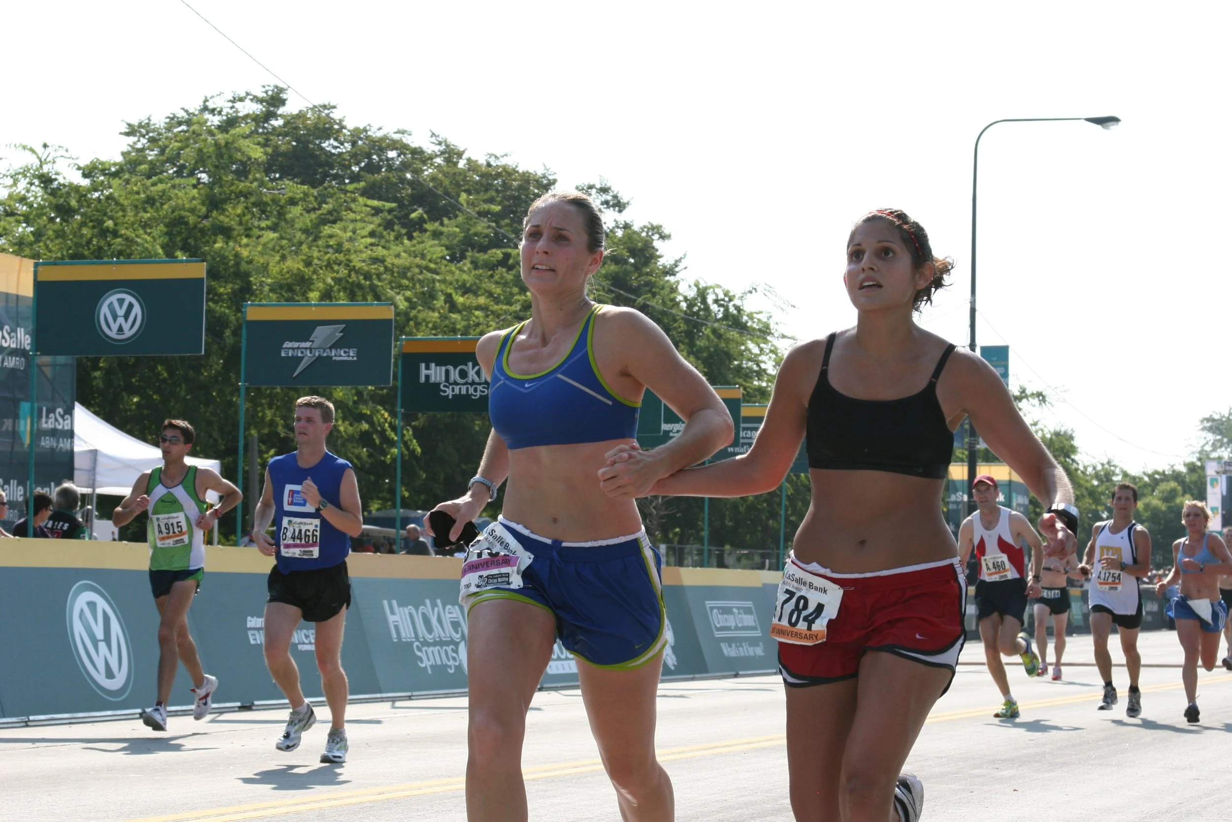 Runners participating in a marathon race on a sunny day, with trees and banners in the background.