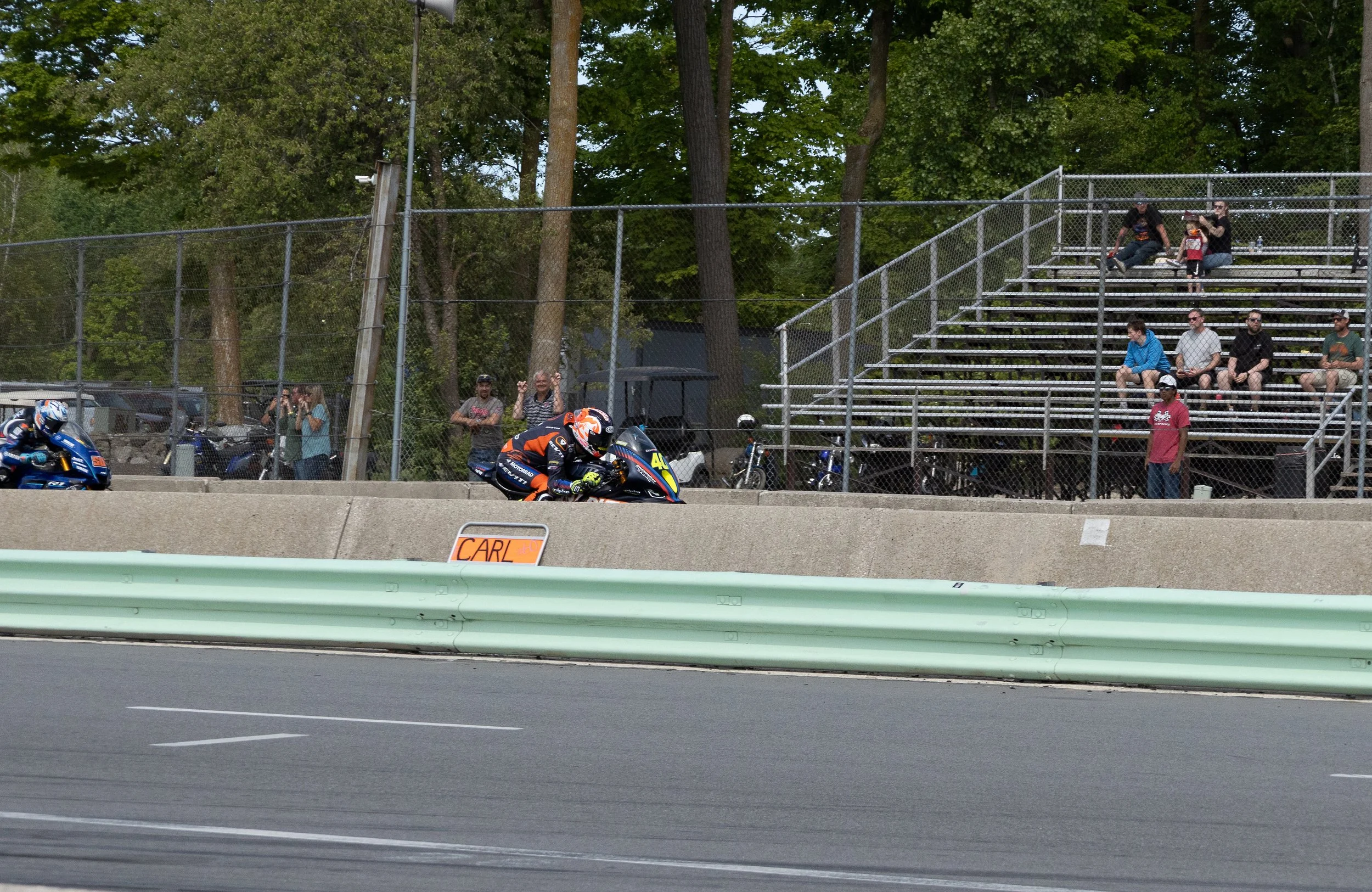 Motorcycle race with two riders on a track, crowd of spectators in bleachers, trees in the background, safety barriers in the foreground.